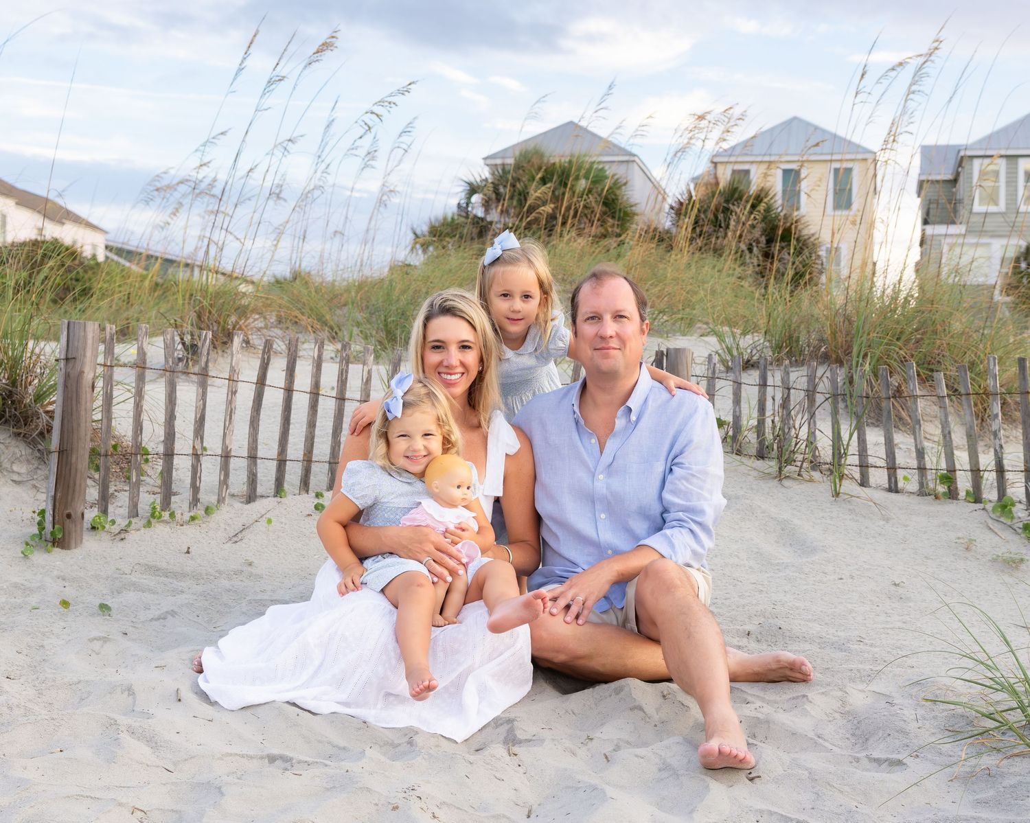 Family of four smiling together on a sandy beach with dune grass and coastal homes in the background on a sunny day.