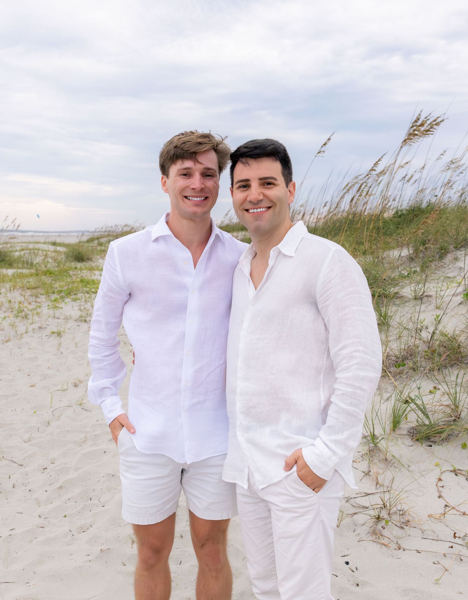 Two men in matching white linen outfits smiling together on a sandy beach with sea oats and cloudy sky.