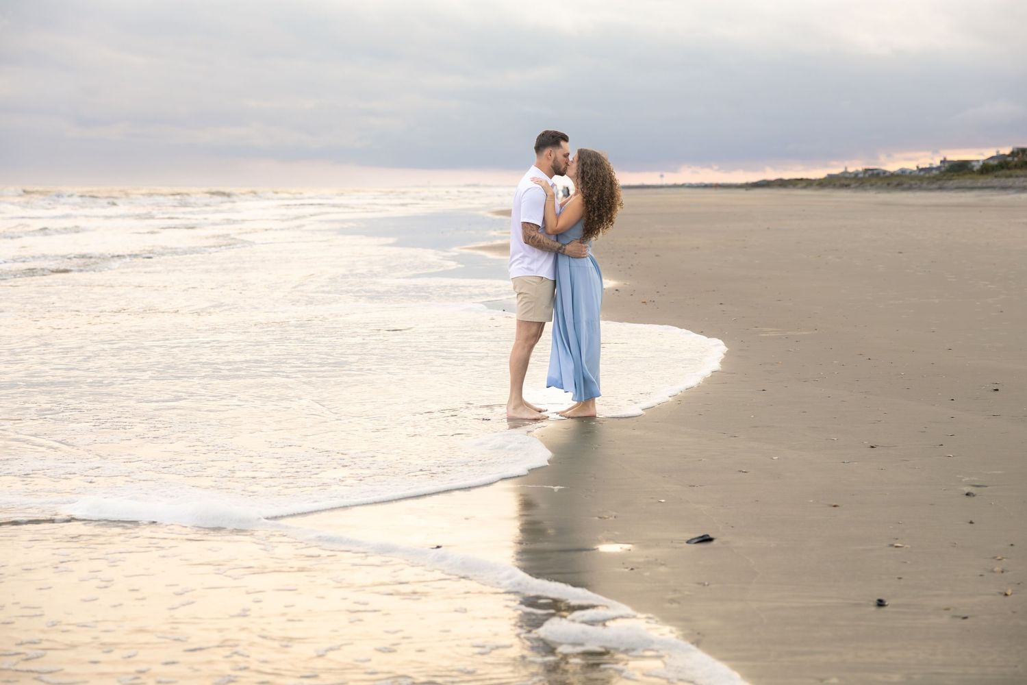 Couple embracing and kissing on a sandy beach at sunset, waves gently washing around their feet.