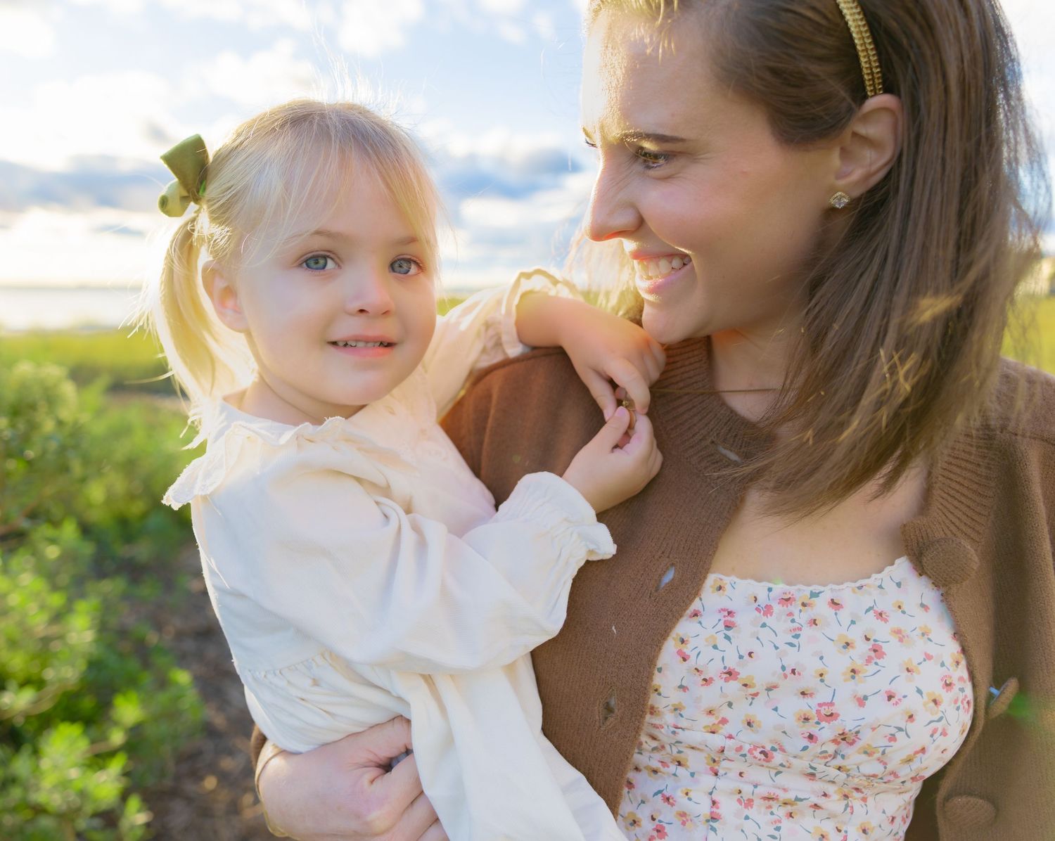 Smiling woman in a brown cardigan and floral top holds a happy toddler girl with a green hair bow outdoors in a sunny grassy field.