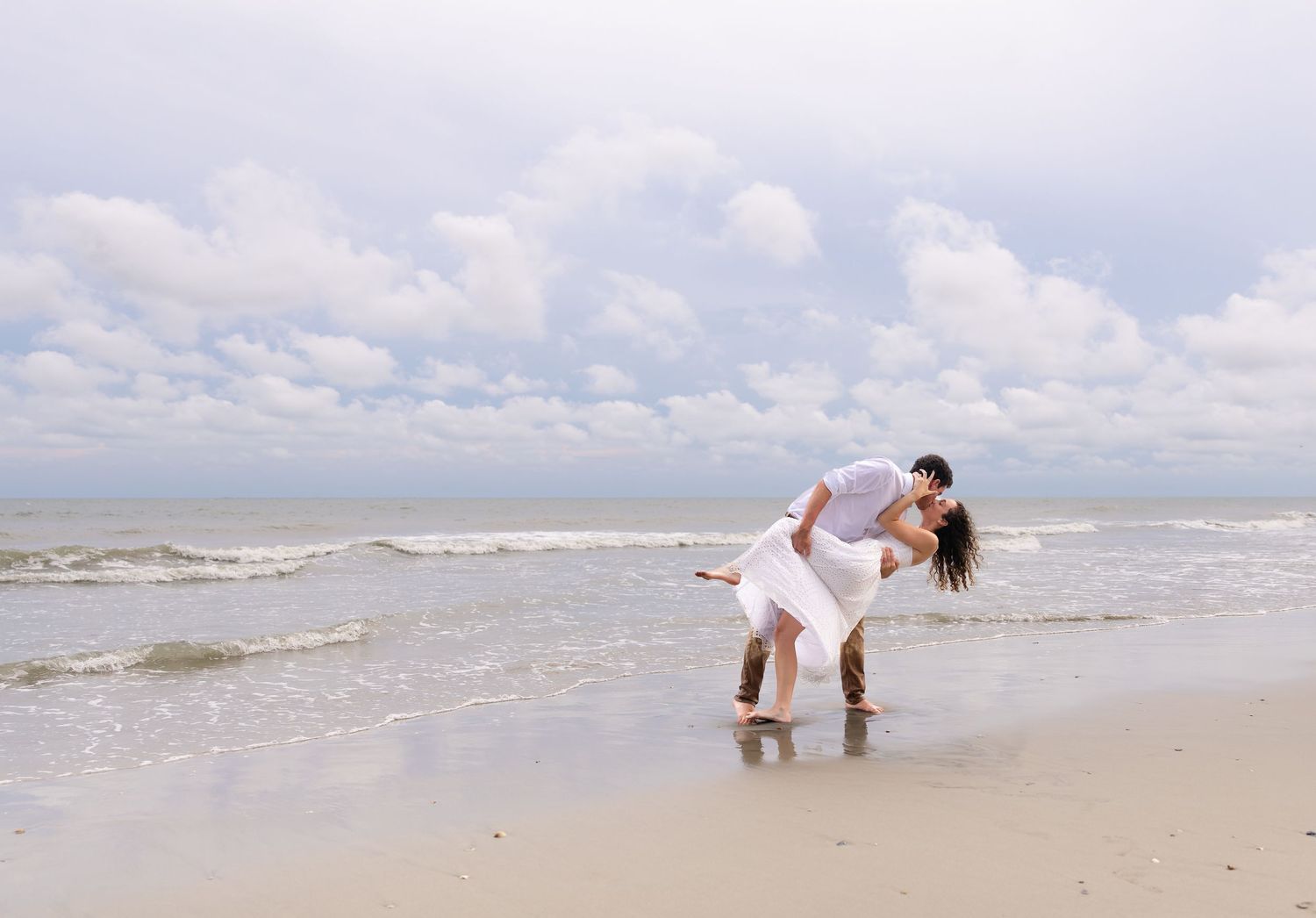 Romantic couple sharing a kiss on a sandy beach with ocean waves and cloudy sky in the background.