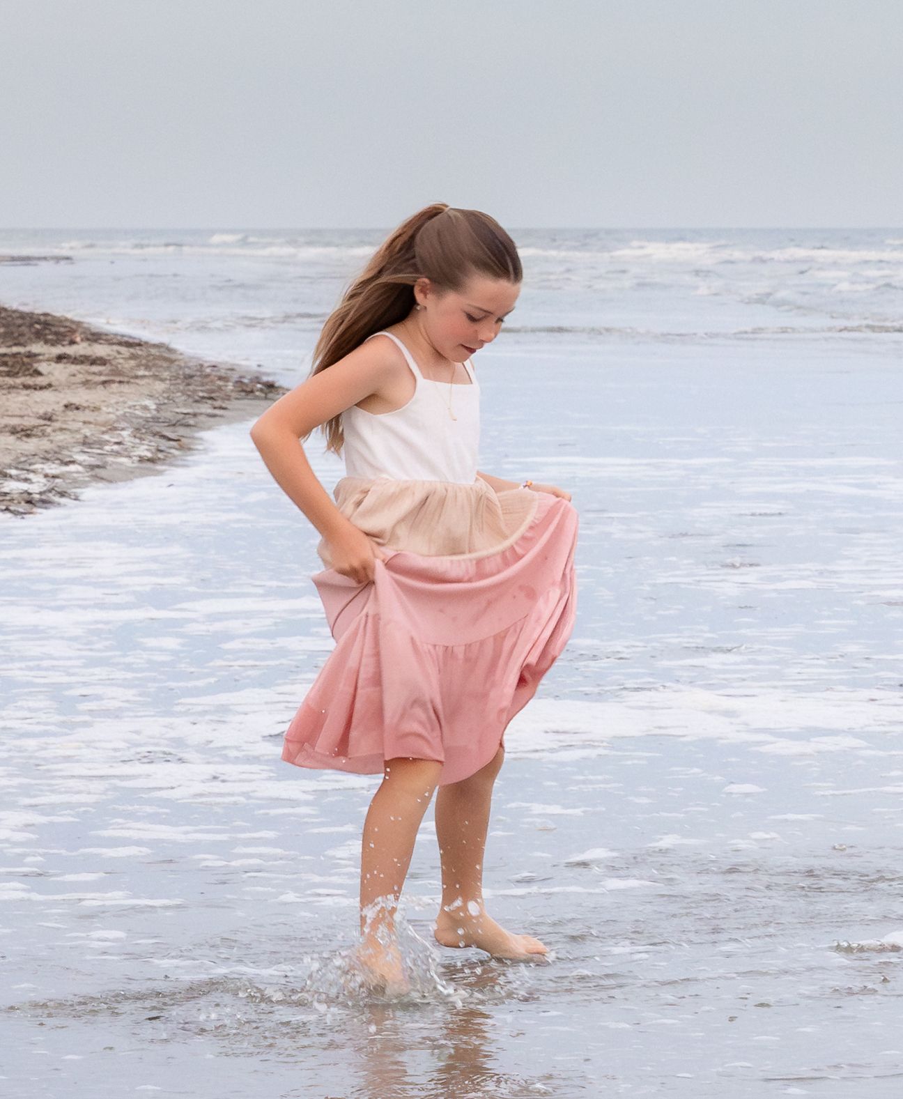 Young girl in pink dress playing in ocean waves at the beach, lifting her skirt as water splashes around her feet.