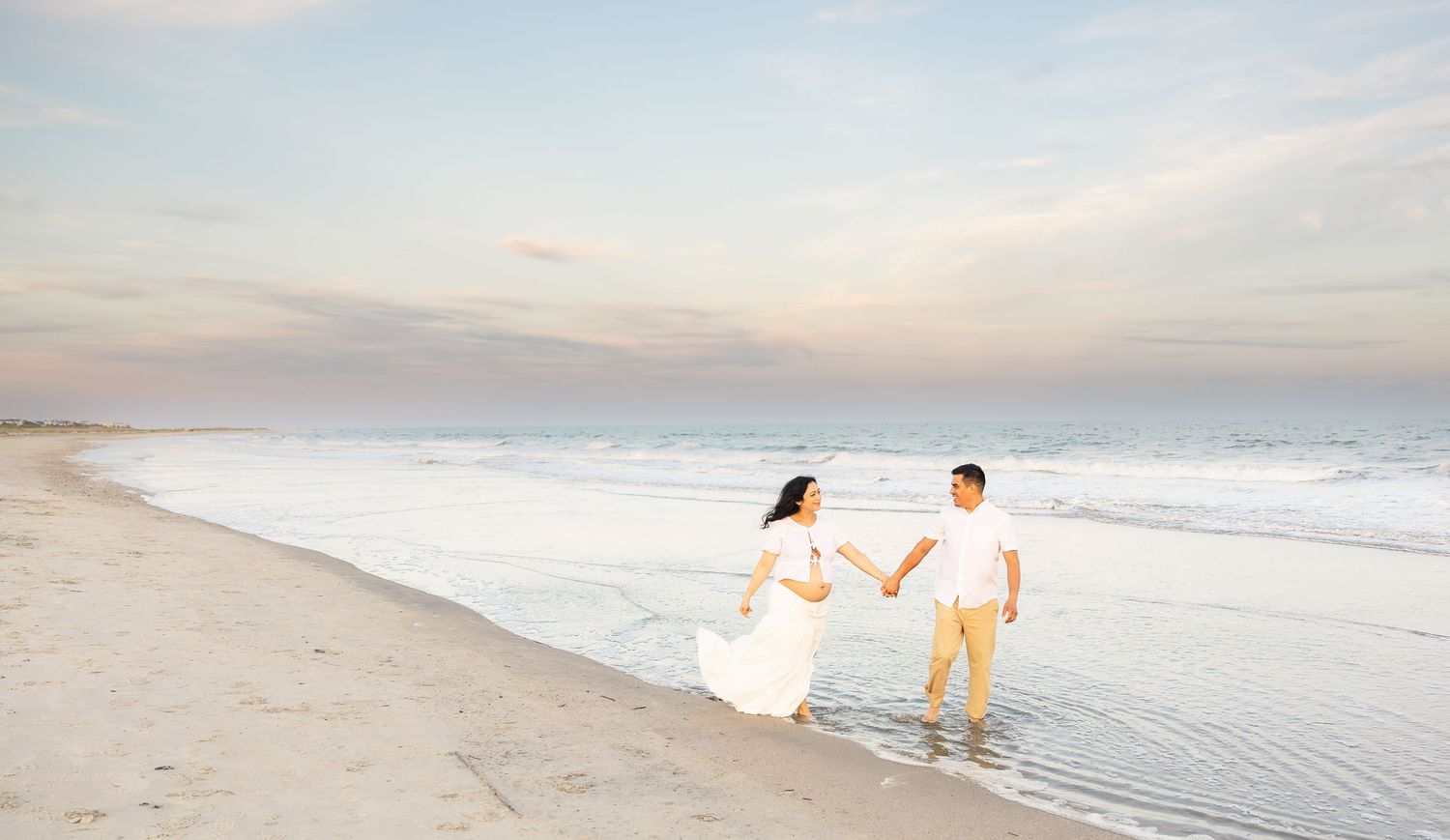 A couple in white attire holds hands while walking along a serene beach at sunset with gentle waves.