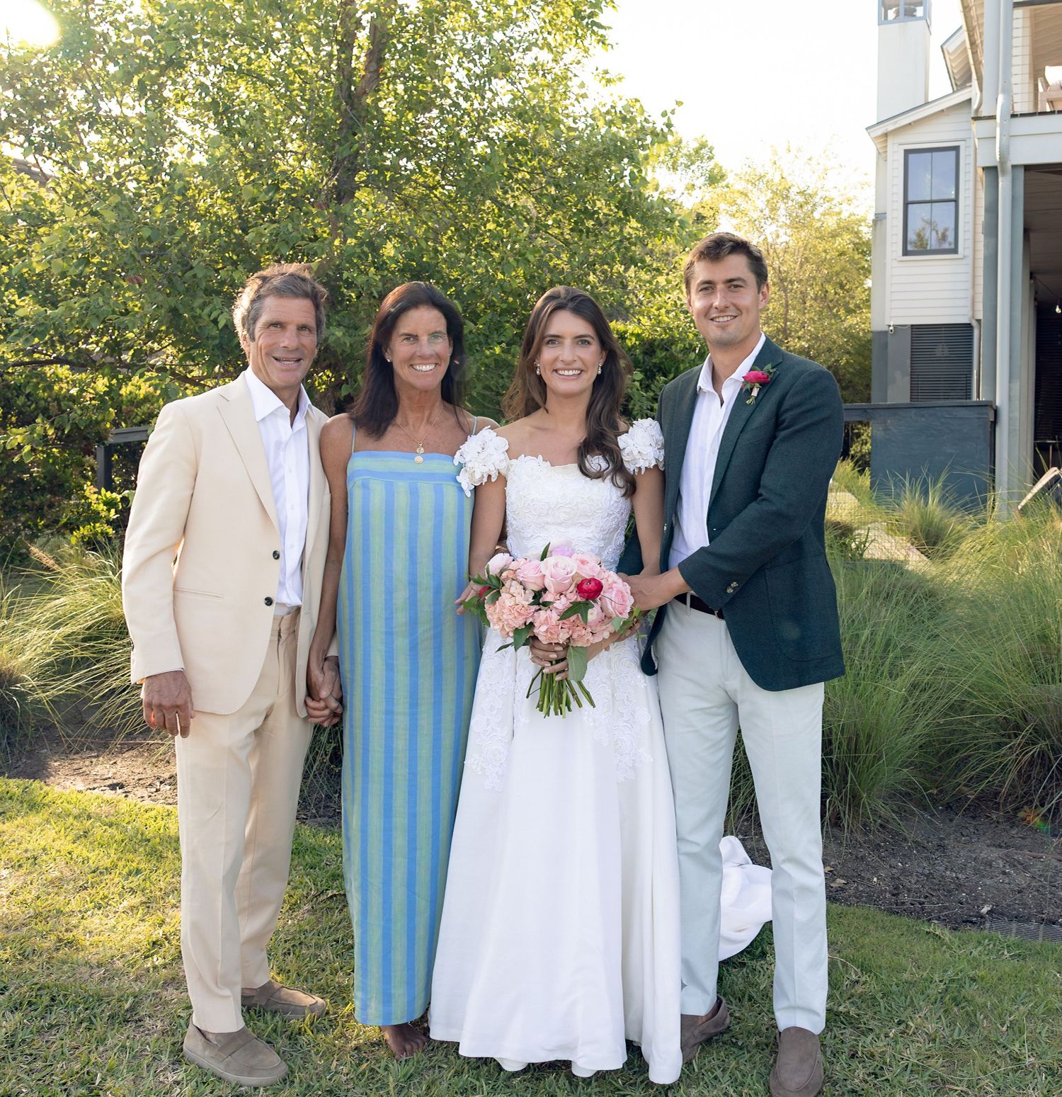Bride in white gown holds pink bouquet posing with three family members outdoors at a backyard wedding celebration.