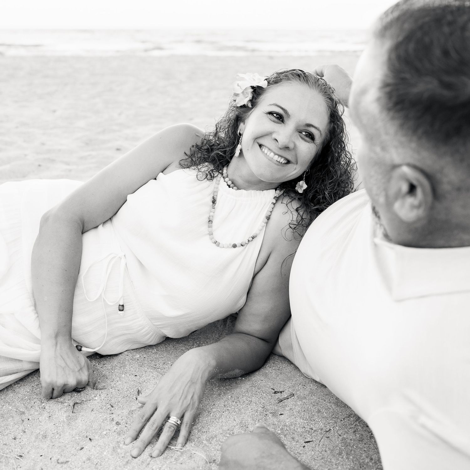 A smiling woman with curly hair and a flower crown lies on beach sand in a white dress, gazing at a man in white.