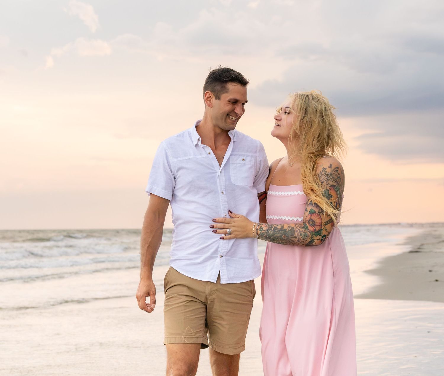 Couple walking on beach at sunset, woman in pink dress with tattoos, man in white shirt and khaki shorts.