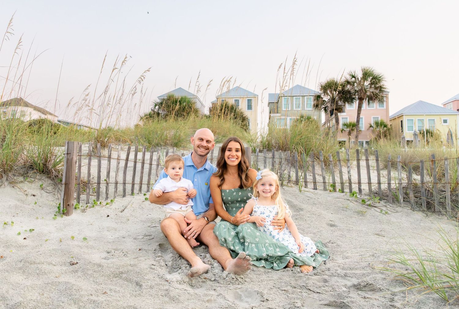 Family of four smiling together on a sandy beach with colorful beach houses and sea oats in the background.