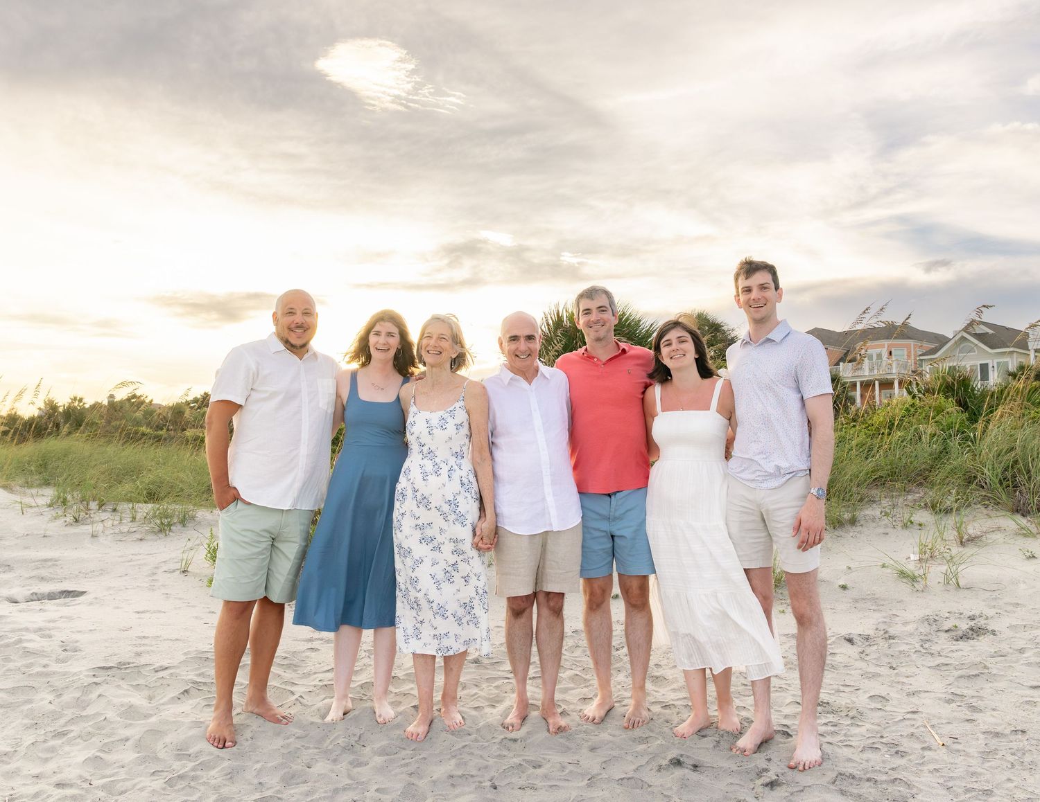 Family of seven posing together on a sandy beach at sunset with coastal homes and dune grass in background.