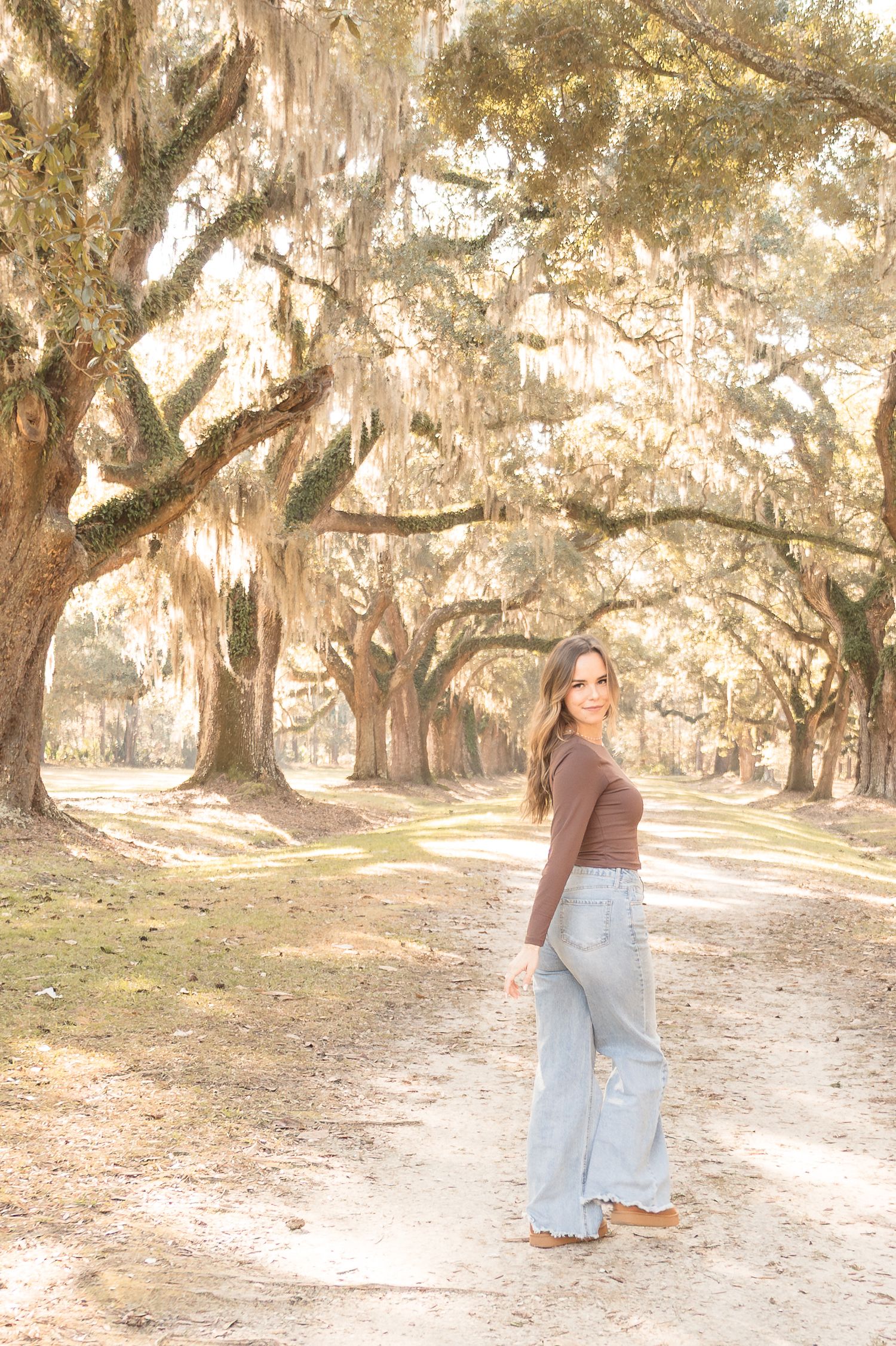 Young woman in wide-leg jeans and brown top poses on a sunlit path lined with moss-draped oak trees.