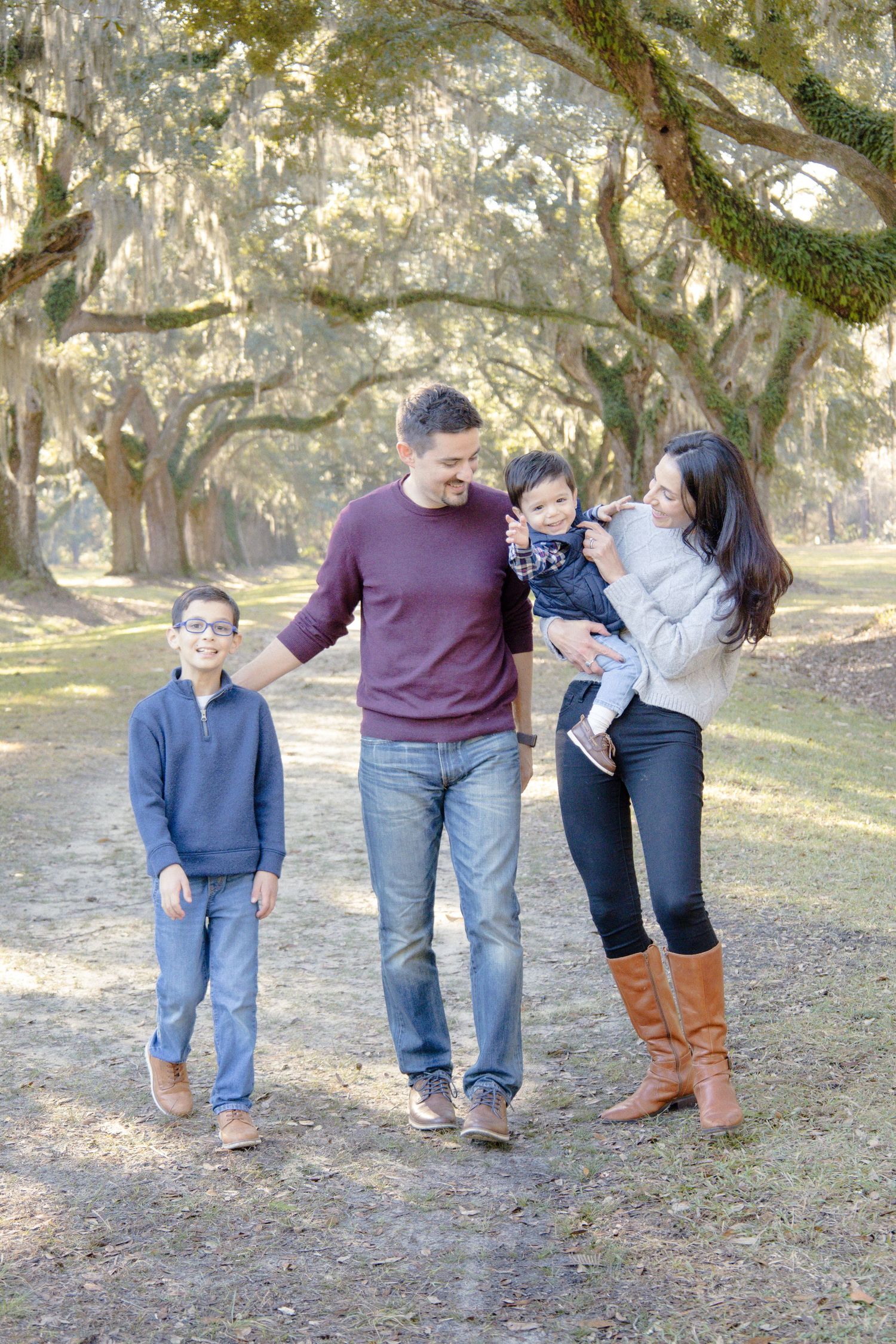 Family of four walking outdoors under moss-covered oak trees, parents smiling at toddler held by mother in brown boots.