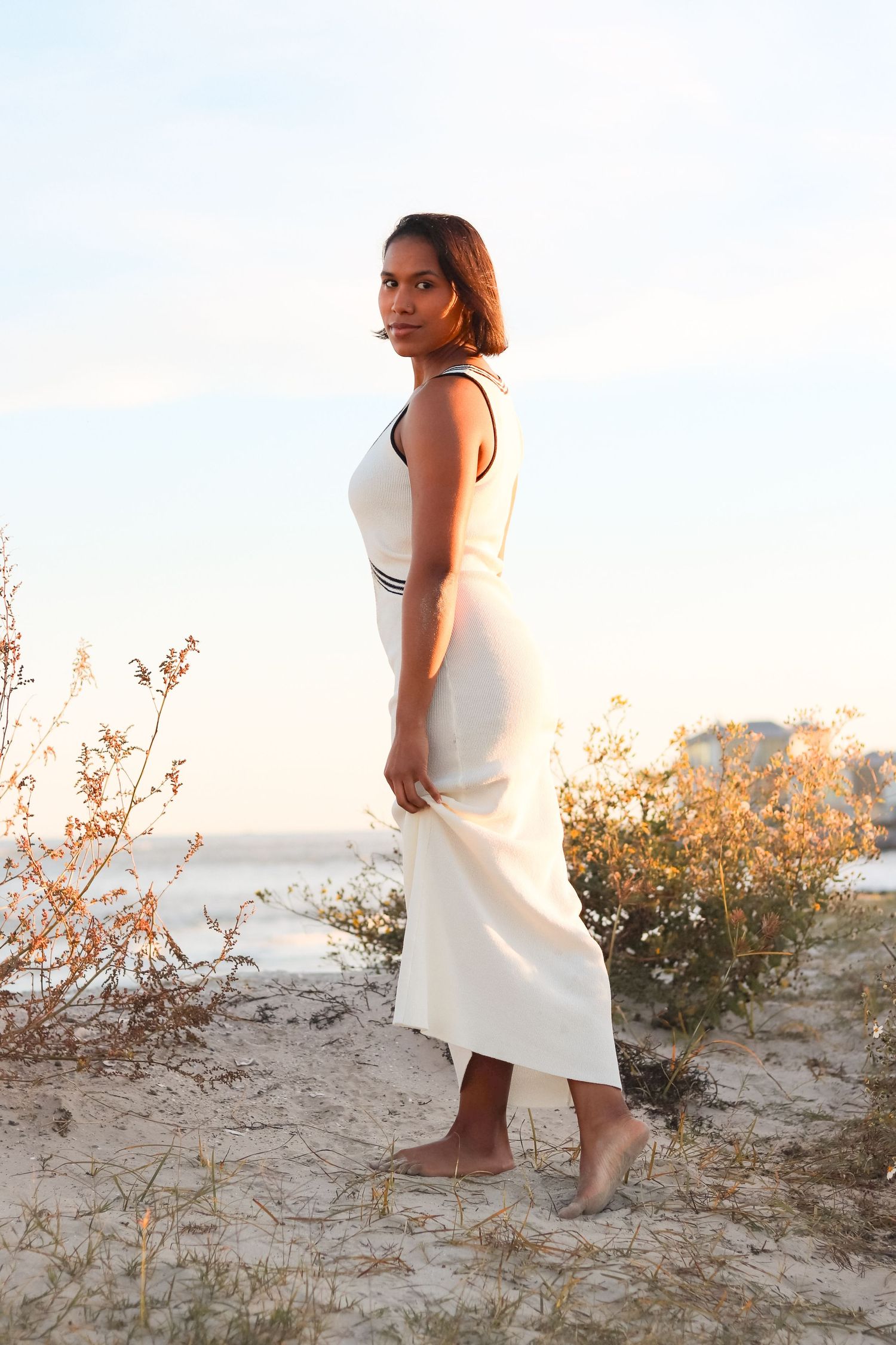 Woman in white maxi dress standing barefoot on sandy beach at golden hour with coastal vegetation.