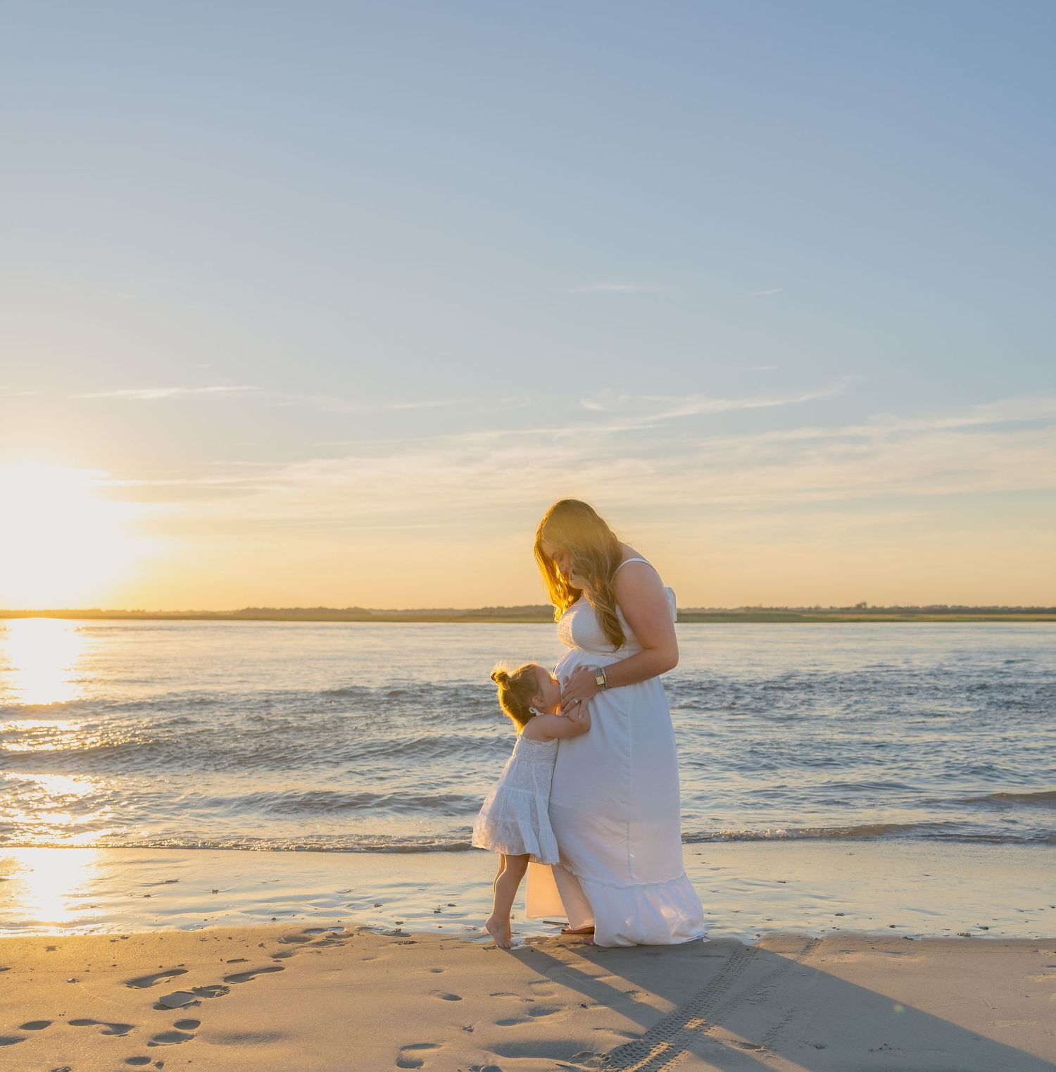 Woman in a white dress embraces a young girl on a sandy beach at sunset, with golden light shimmering over calm ocean waves.