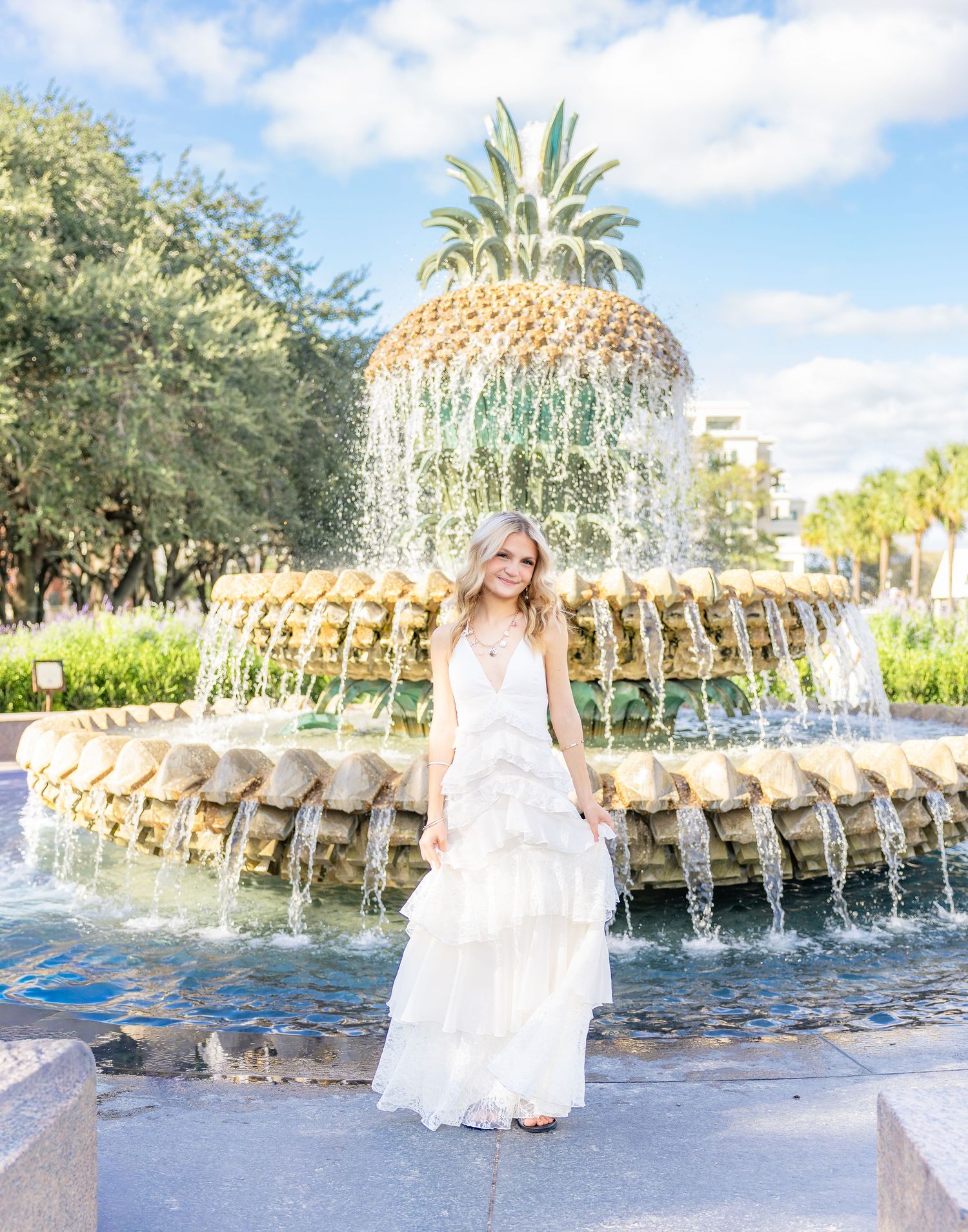Young woman in a flowing white tiered dress poses in front of the pineapple fountain with cascading water on a sunny day.