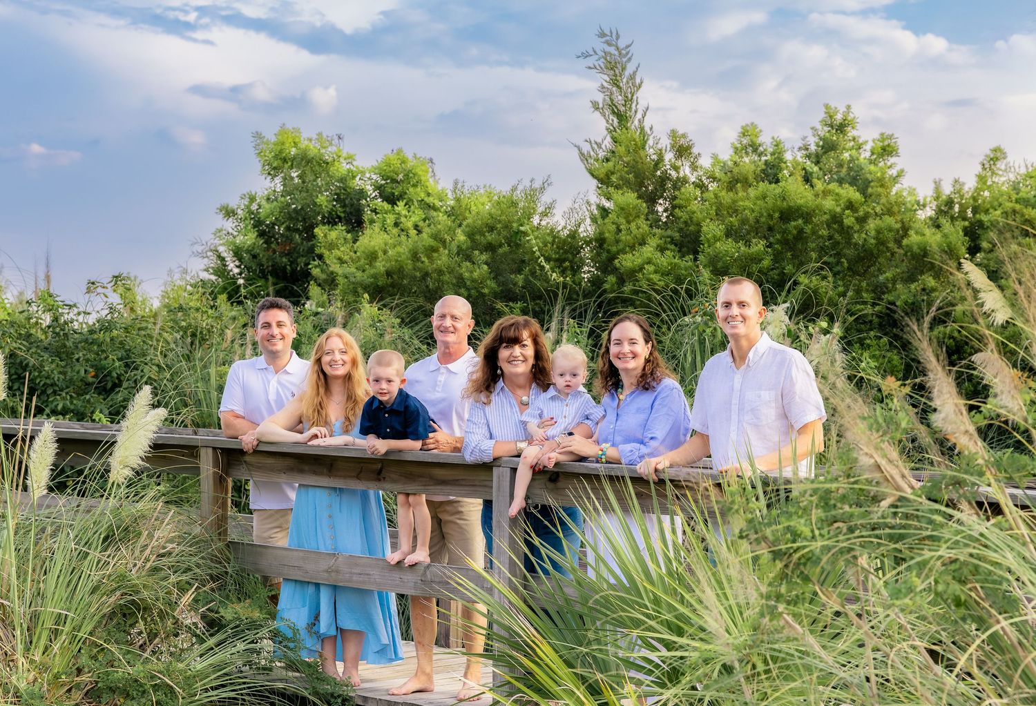 Smiling family of eight posing together on a wooden boardwalk surrounded by tall beach grasses and green trees under a blue sky.
