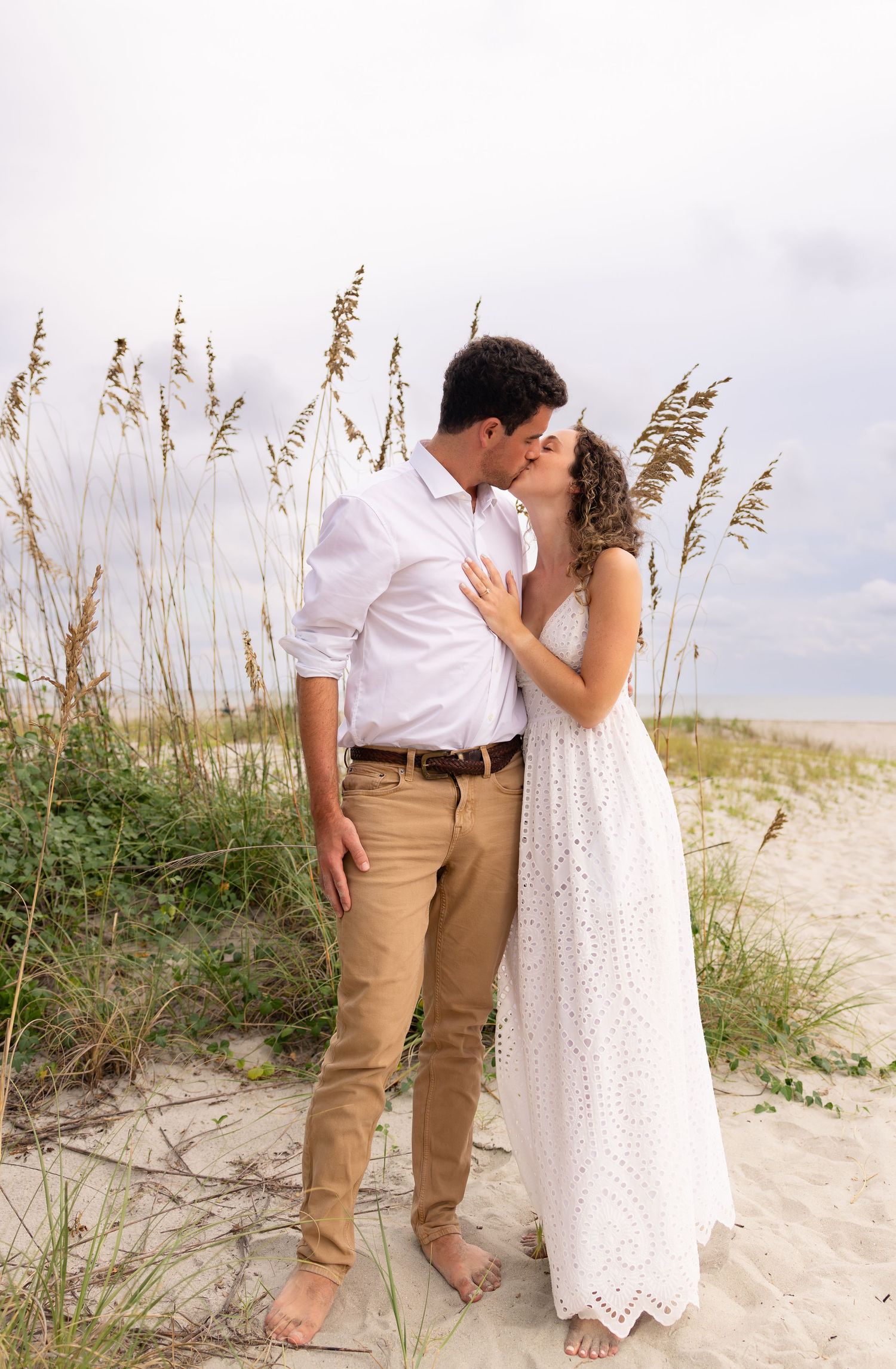 Barefoot couple in white and khaki kissing on a sandy beach surrounded by tall sea oats under a soft cloudy sky.