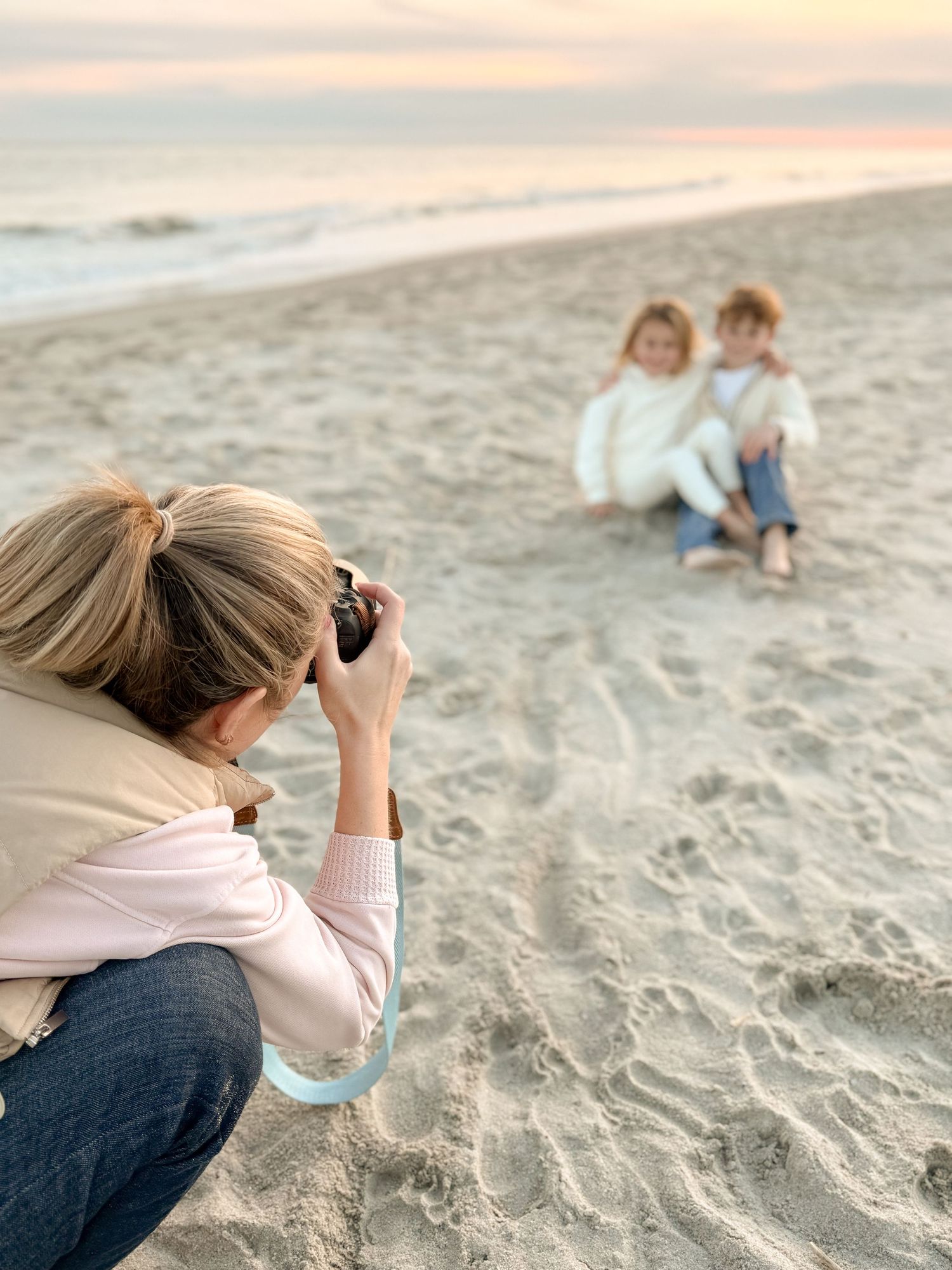Photographer crouching on sandy beach taking pictures during sunset with soft pastel sky in background.