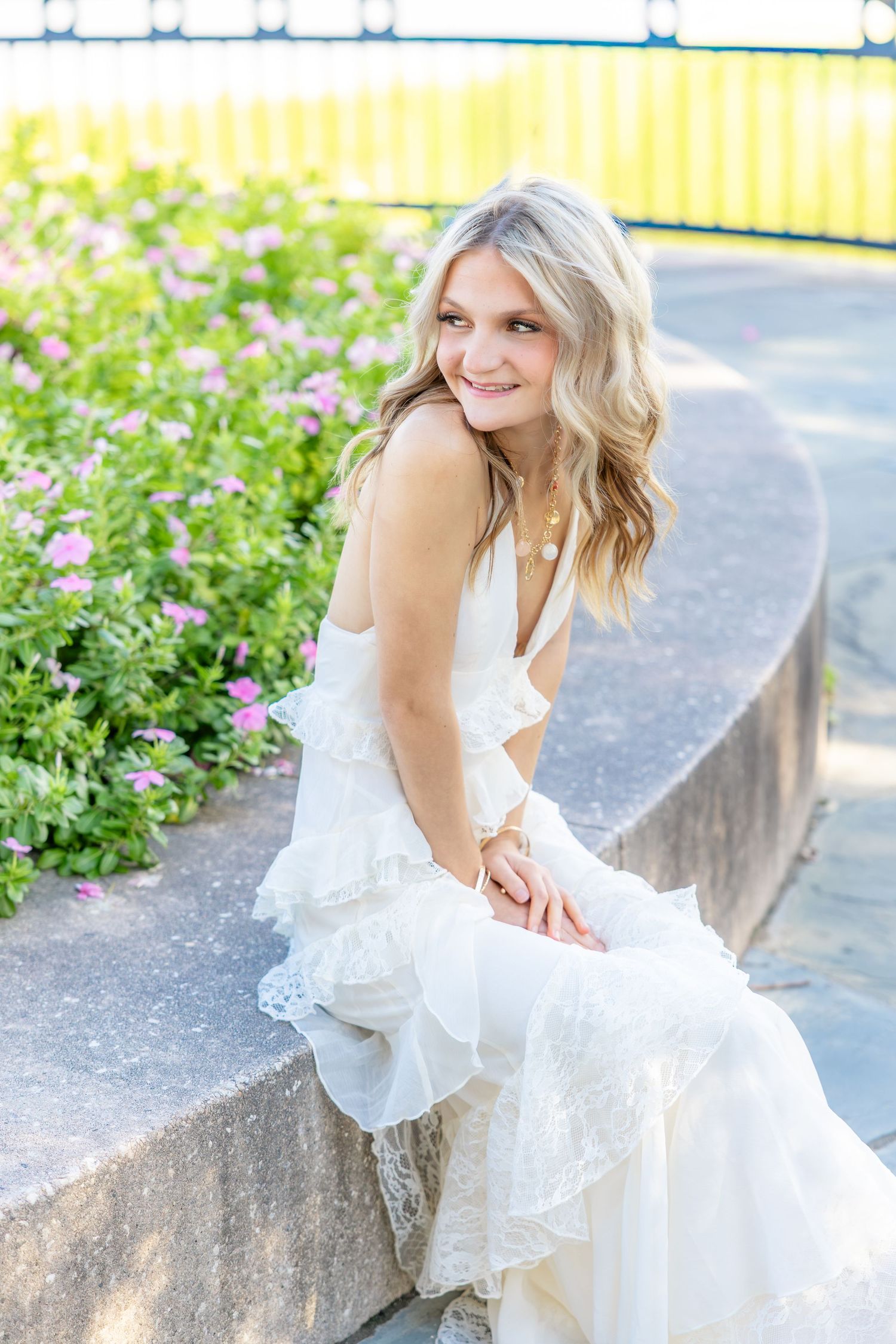 Smiling woman with wavy blonde hair in a white ruffled lace dress sits on a curved stone ledge beside pink flowers on a sunny day.