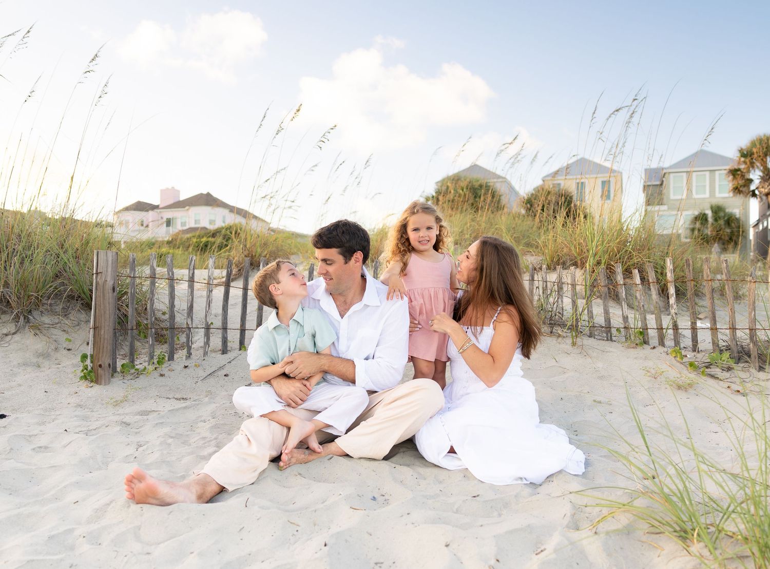 Happy family of four sitting together on sandy beach dunes near a wooden fence on a sunny day.