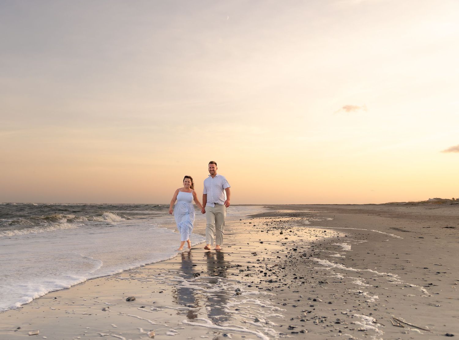Couple holding hands and walking along the wet sandy shoreline of a beach at sunset, with gentle waves and a warm pastel sky behind them.