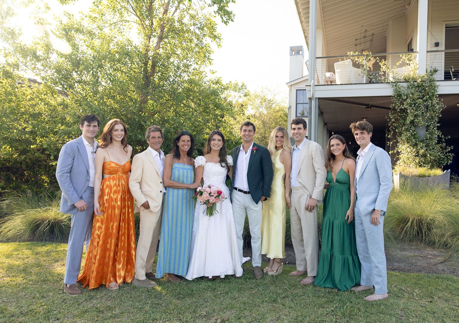 Wedding party group portrait with bride and groom surrounded by guests on sunny outdoor lawn.