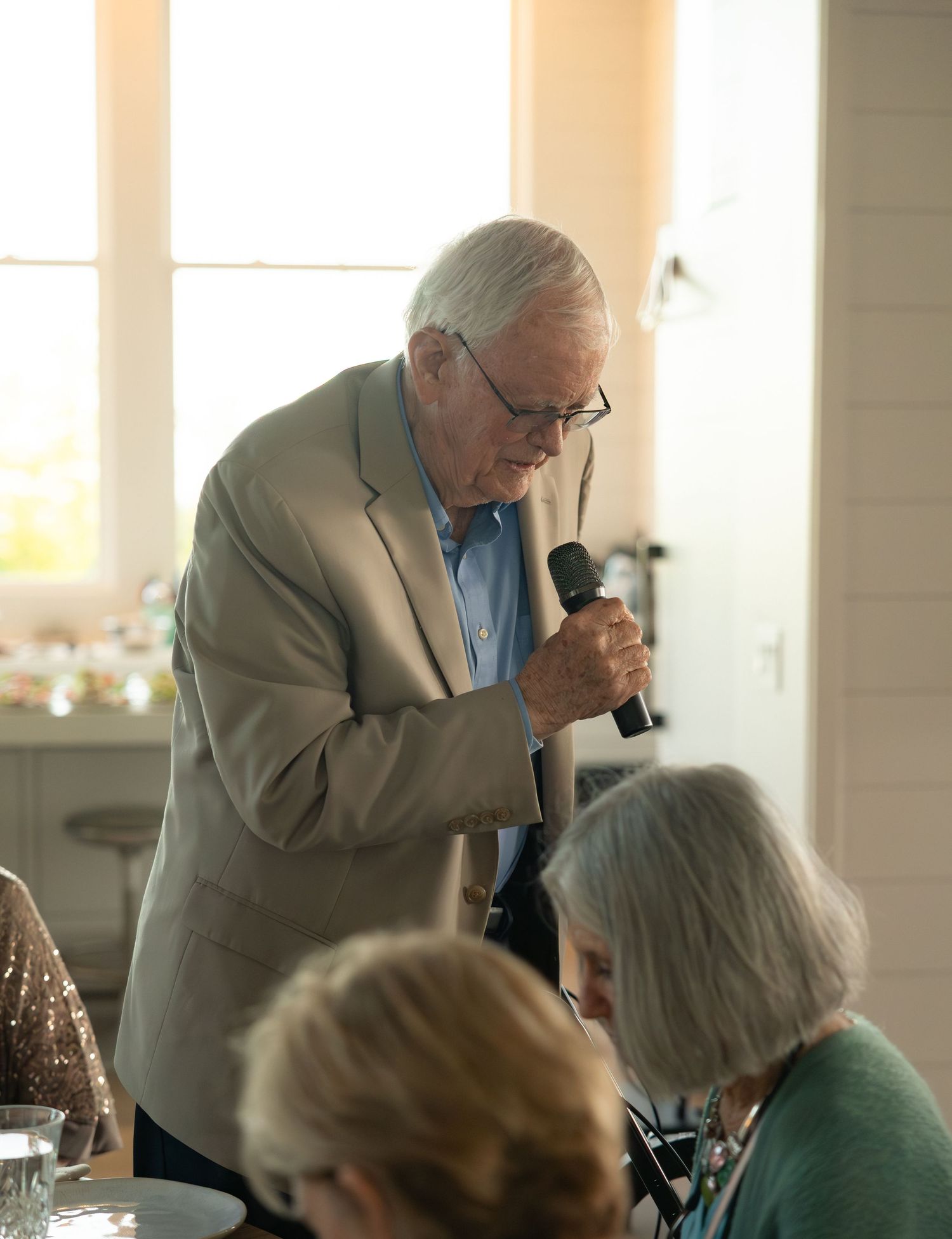 Elderly man in beige suit giving speech with microphone at wedding reception dinner.