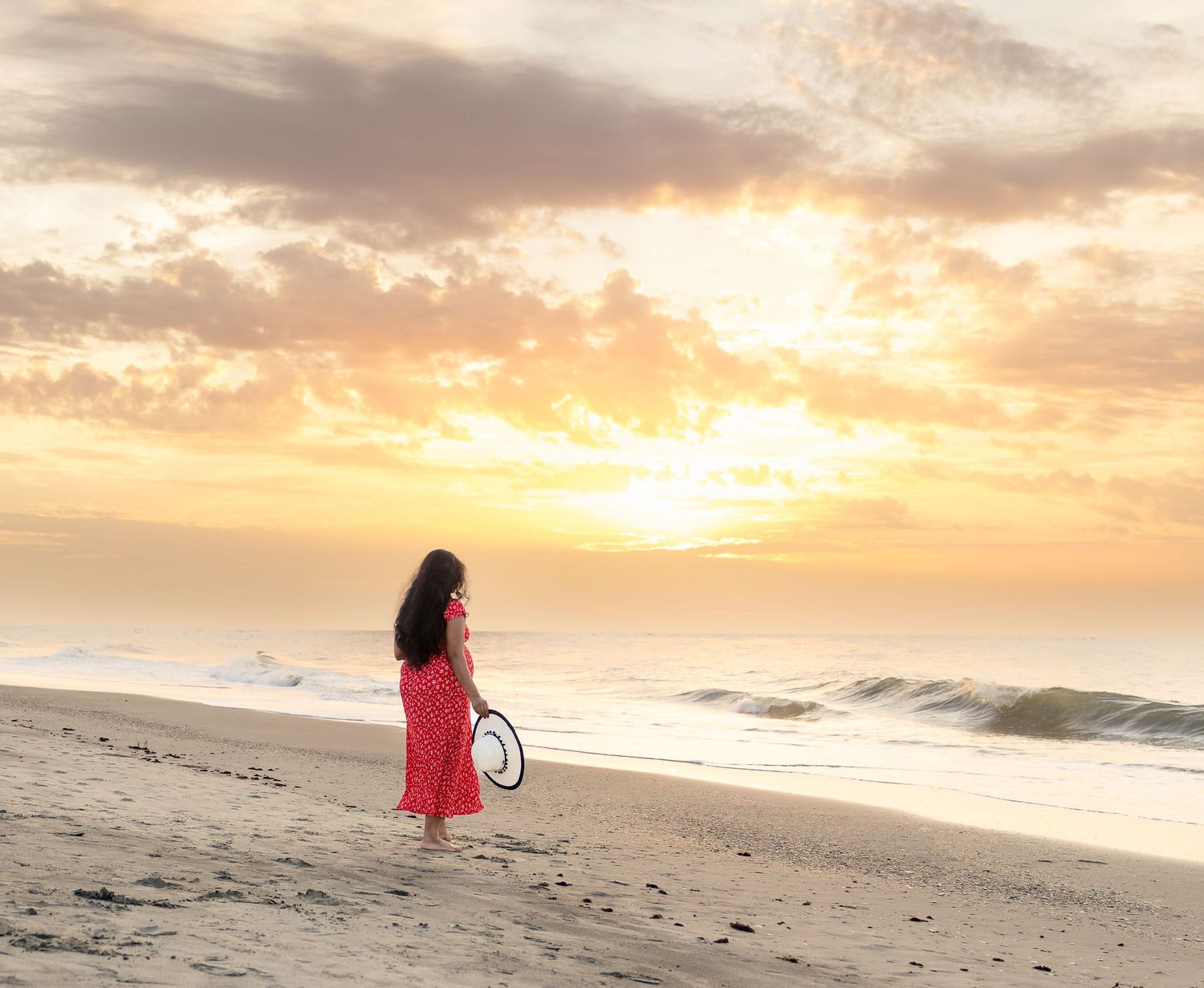 Woman in red dress holding a tennis racket stands on a sandy beach watching a golden sunset over the ocean.
