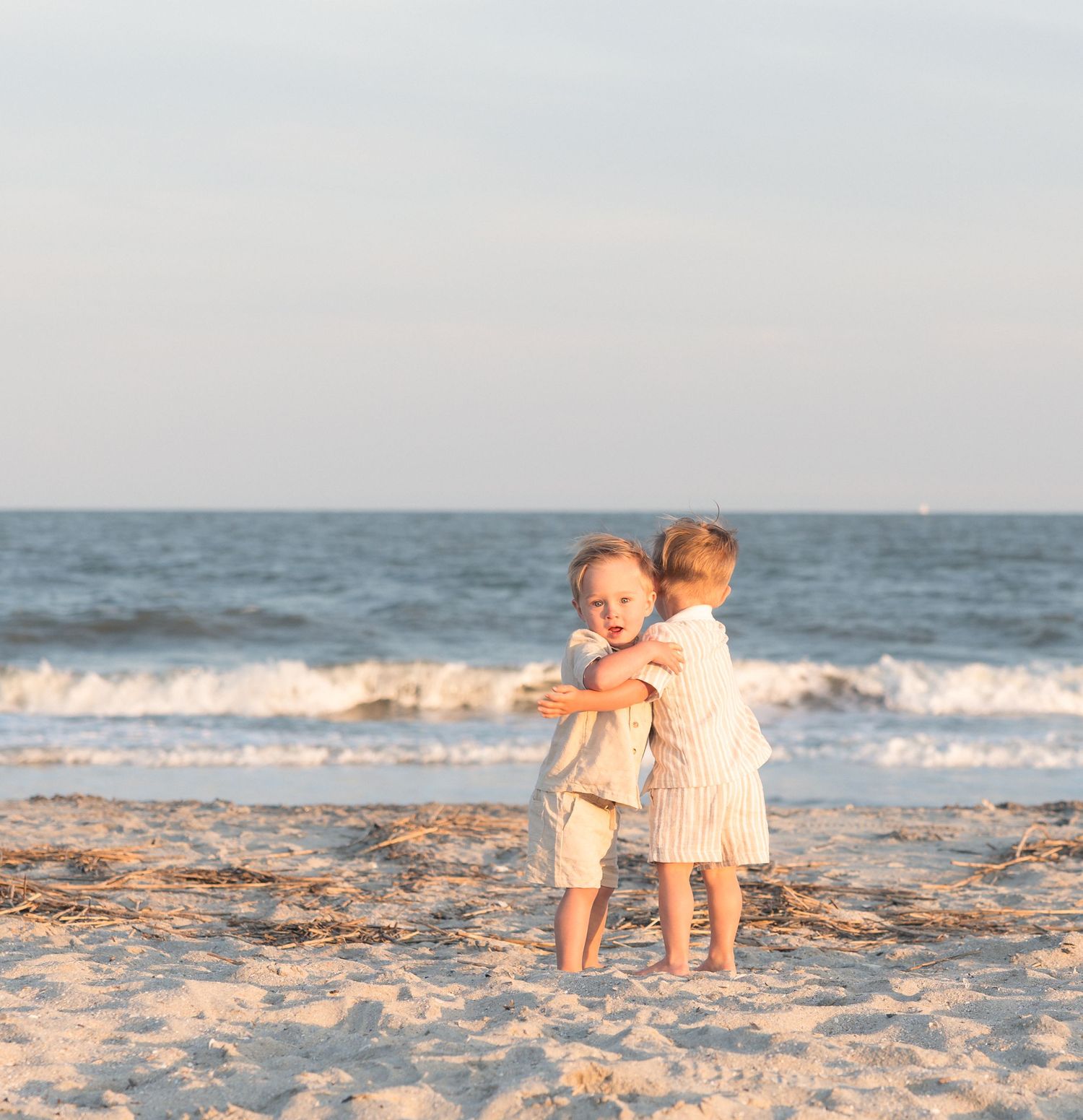 Two young siblings share a sweet hug on a sunlit beach with ocean waves in the background.