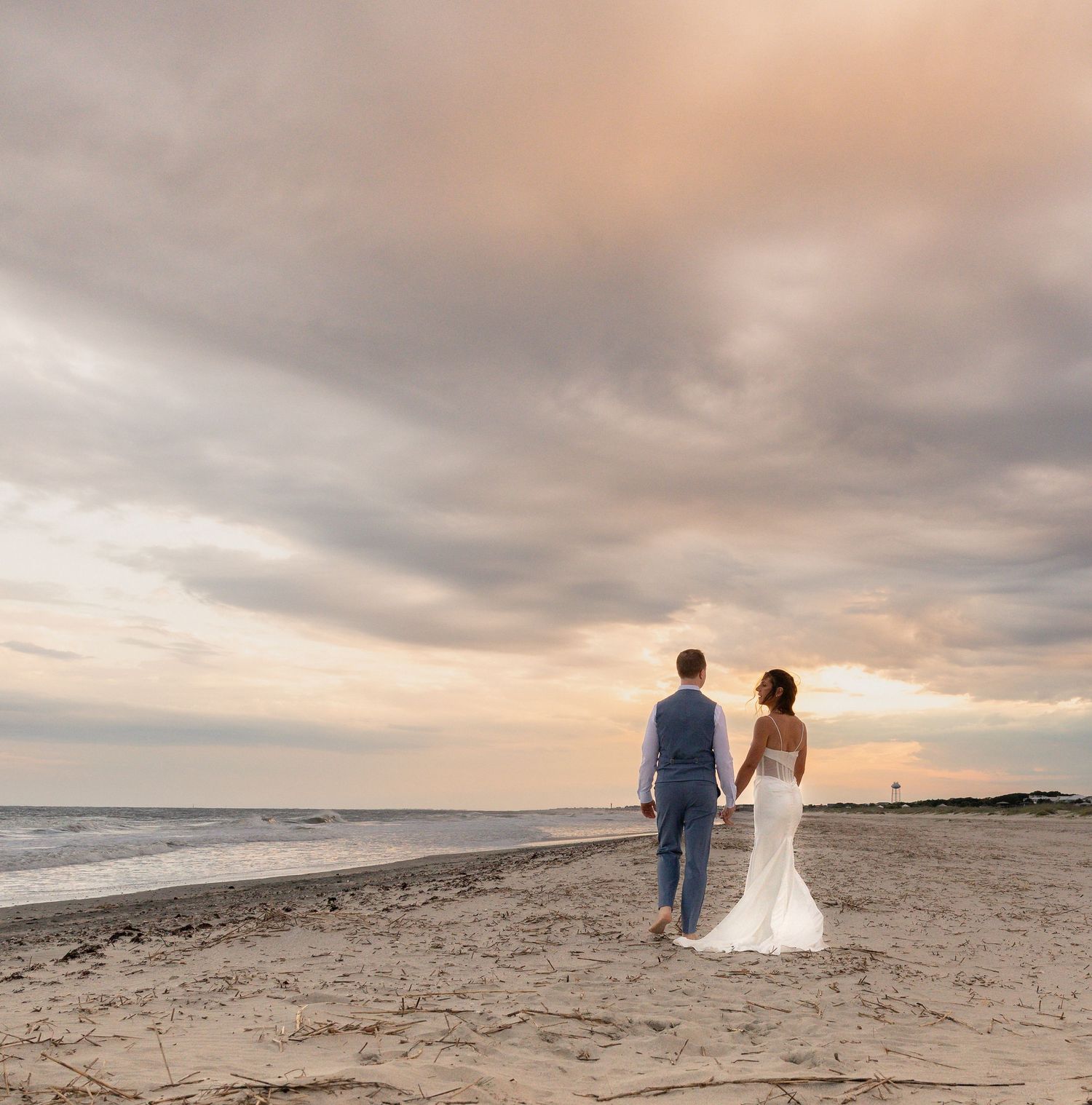 A bride and groom hold hands while walking along a sandy beach at sunset under a dramatic cloudy sky.
