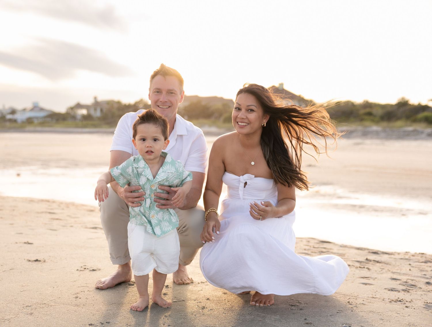 Family of three posing on a sandy beach at golden hour, parents smiling with toddler in tropical shirt.