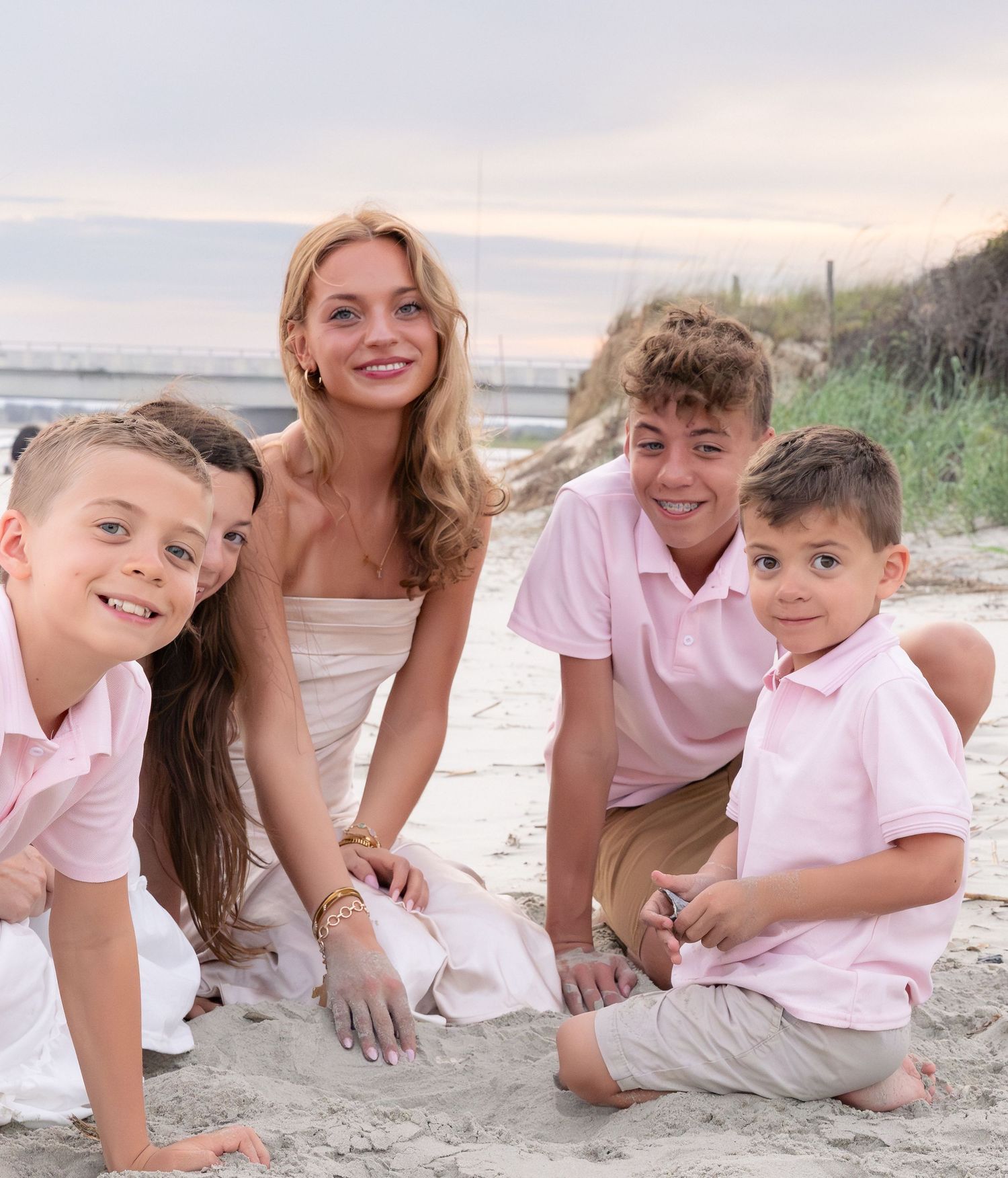 A woman and three boys in pink shirts smile together on a sandy beach at sunset.