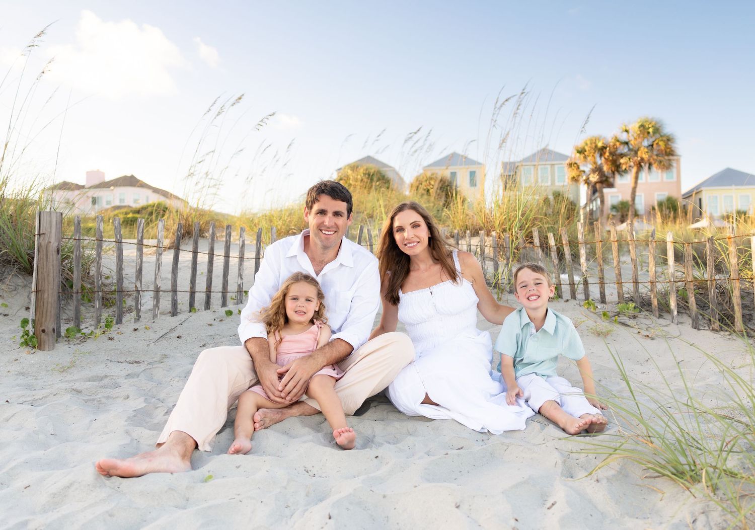 Happy family of four sitting on sandy beach dunes with sea oats and colorful beach houses in background.