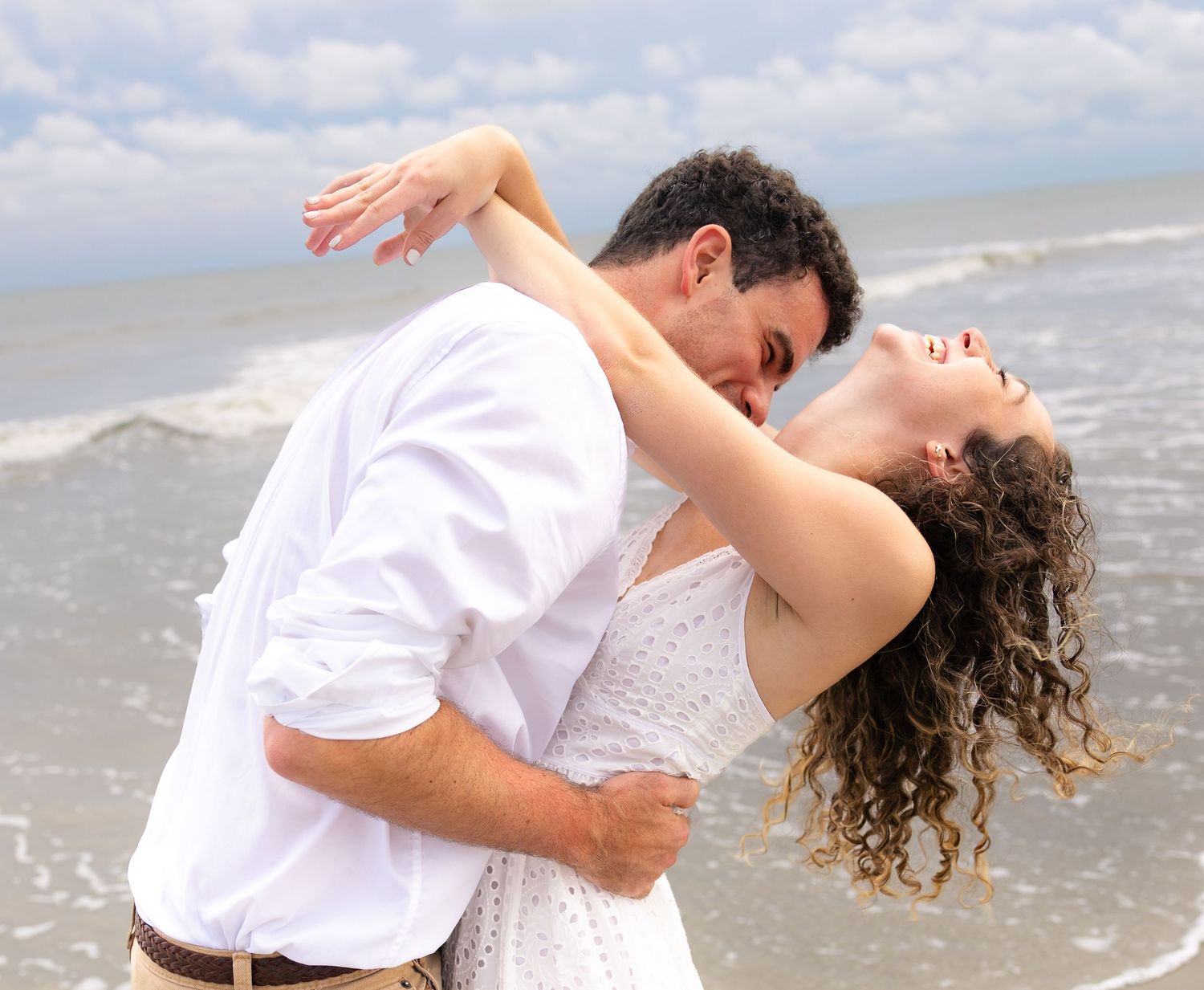 Romantic couple kissing and dancing on a beach, dressed in white, with ocean waves in the background.