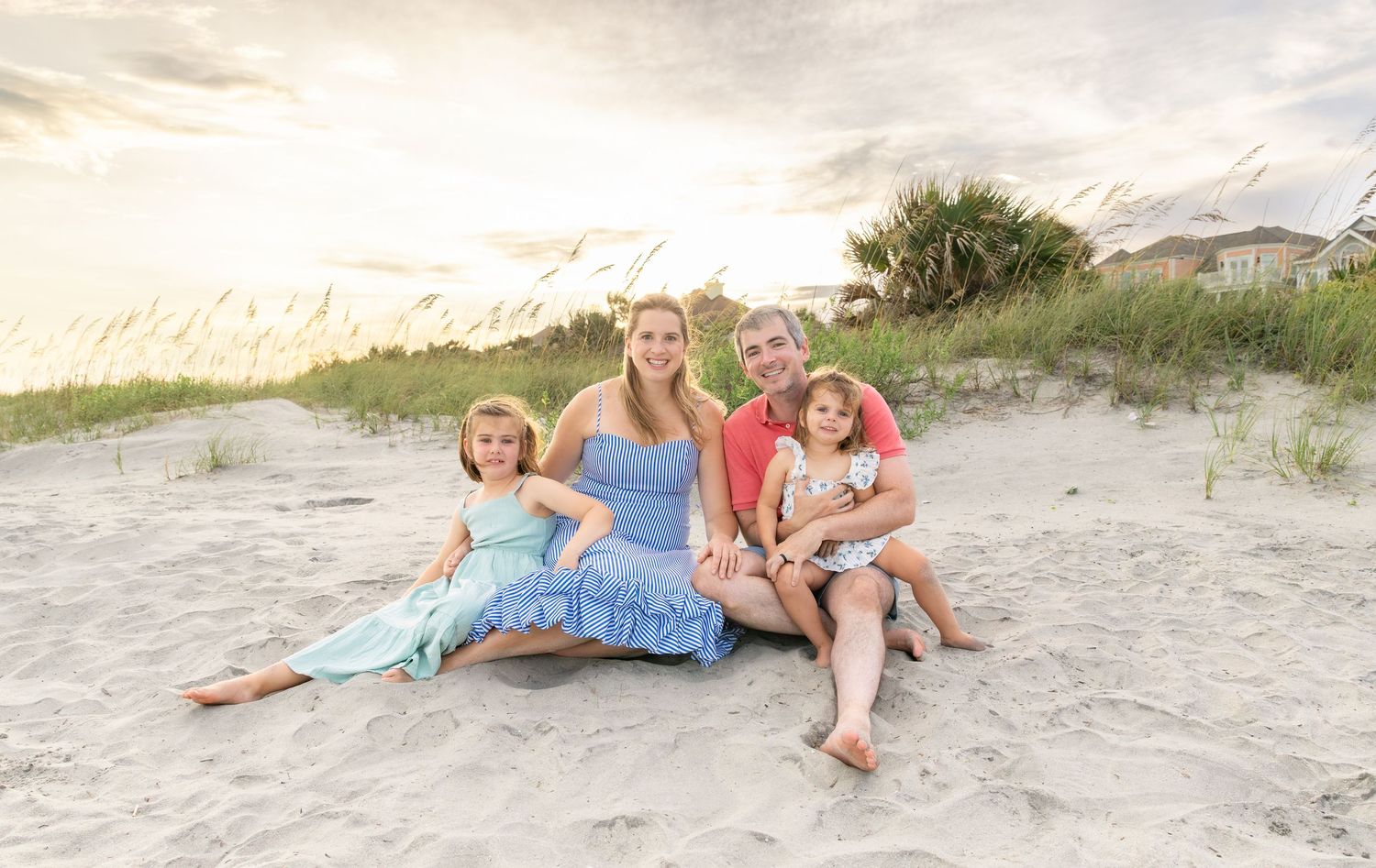 Family of four smiling together on a sandy beach at sunset, with sea oats and pastel sky in the background.