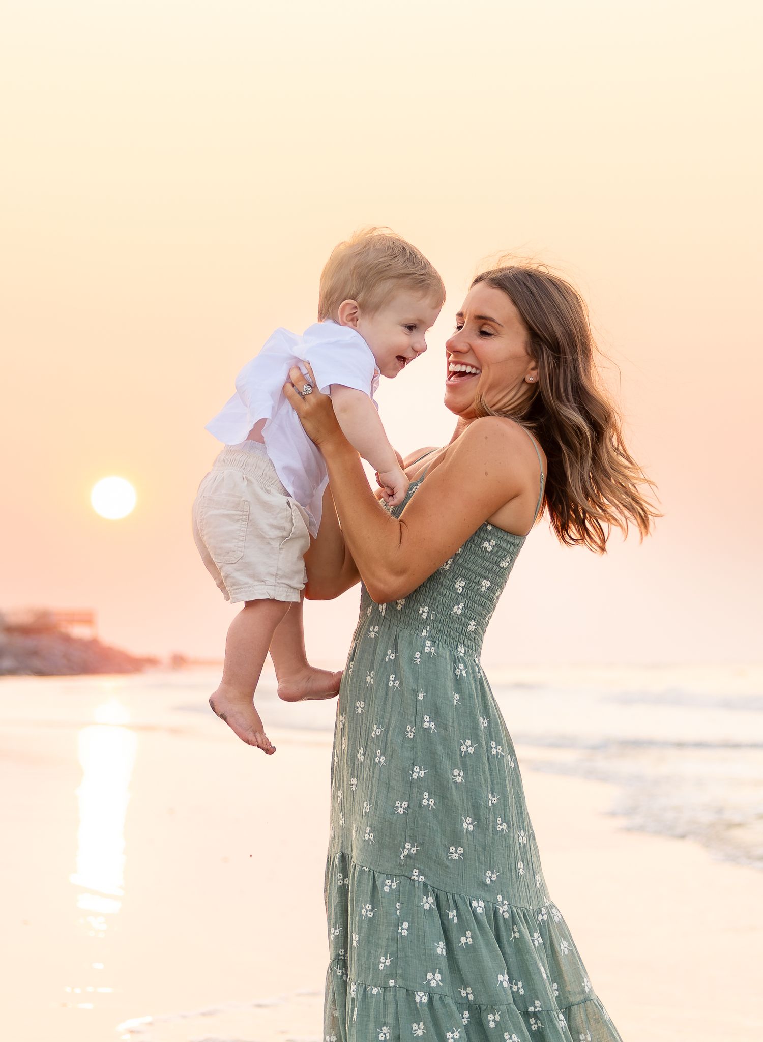 A joyful mother in a green floral dress lifts her laughing baby boy at sunset on a beach.