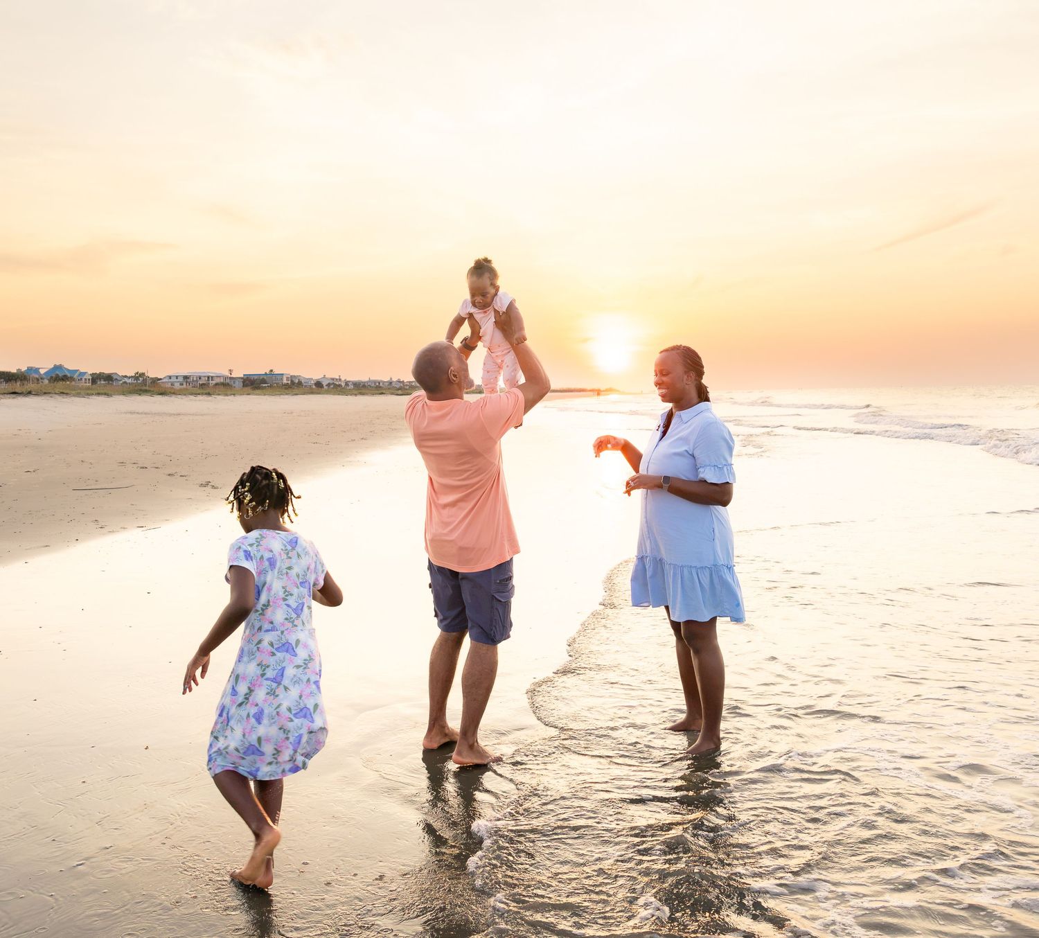 Family enjoying a beach sunset, father lifting baby while mother and daughter play in the waves on a golden evening.