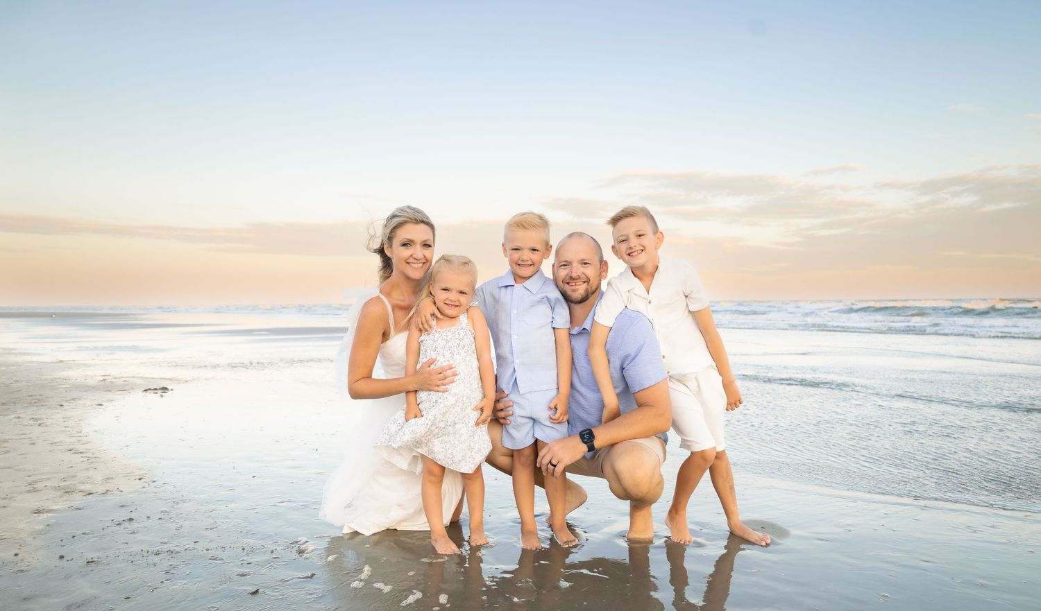 Happy family of five posing together on a sandy beach at sunset, wearing white and blue outfits.