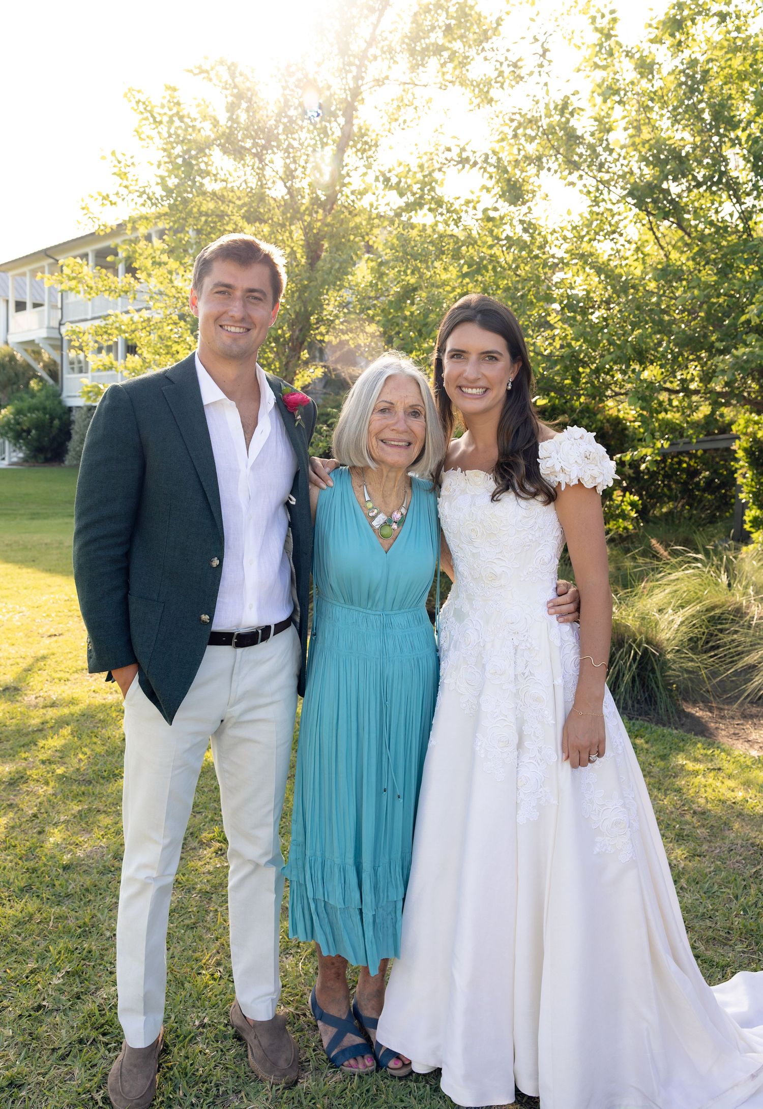 A bride in white gown poses with groom in green blazer and older woman in teal dress at outdoor wedding.