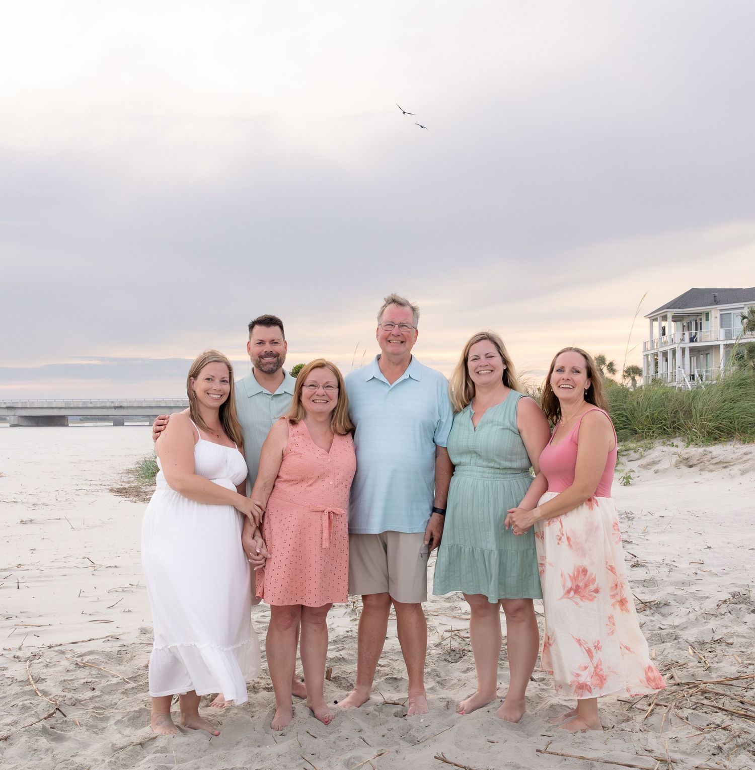 Family of six smiling together on a sandy beach at sunset, wearing pastel summer outfits.