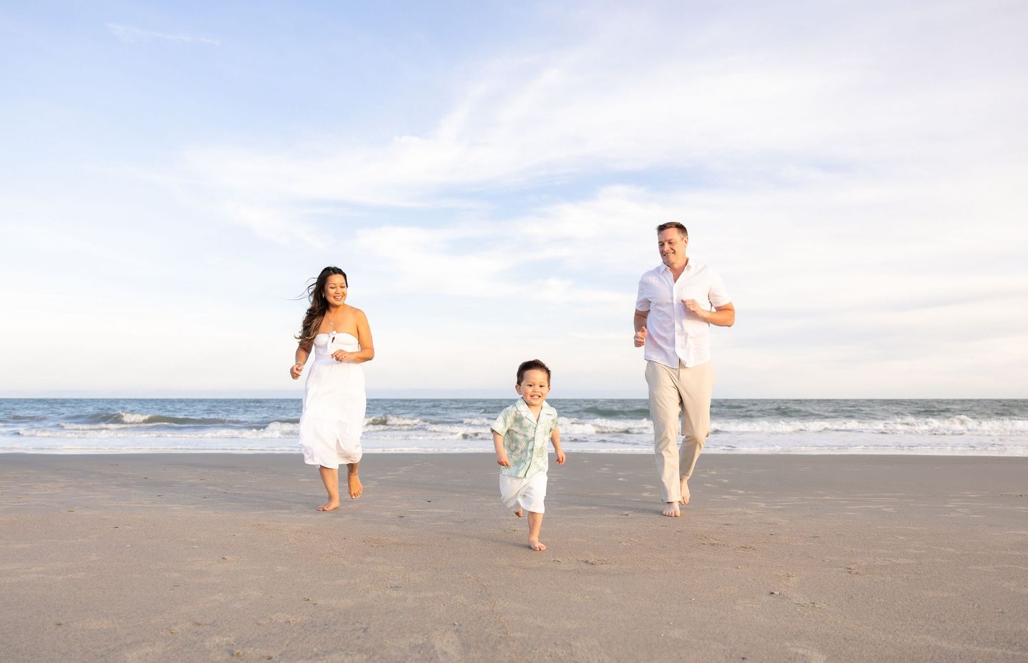Family of three running joyfully on a sunny beach, parents in white outfits with young child between them.