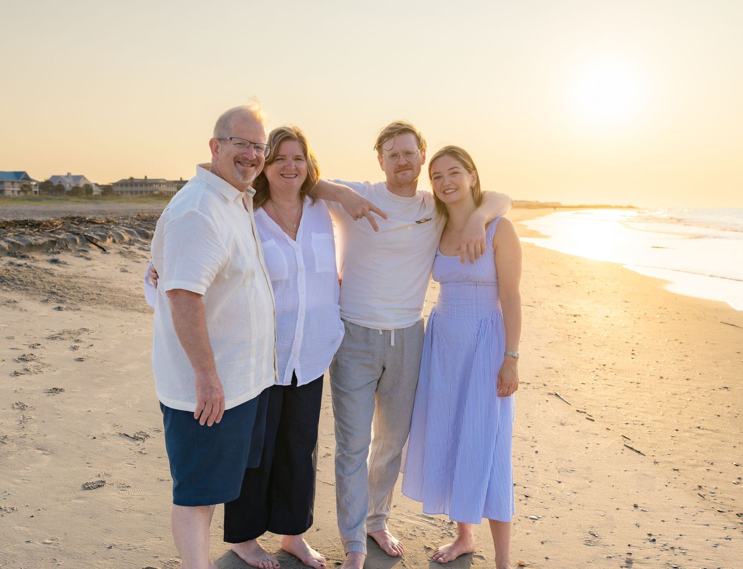 Four people smiling together on a sandy beach at golden sunset hour, posing for a family portrait by the ocean shore.