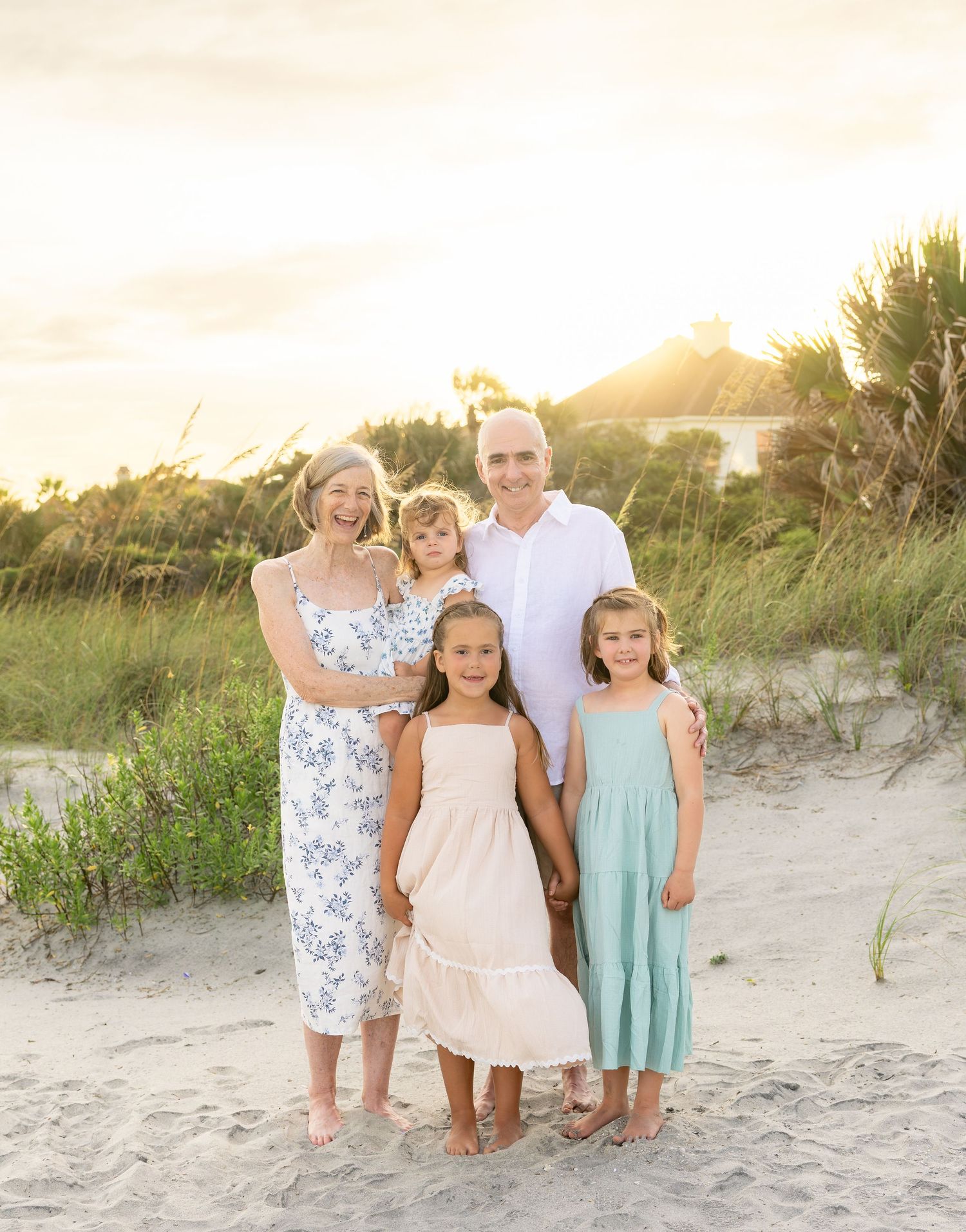 Family of five posing on sandy beach at golden hour, two adults and three girls in summer dresses smiling.