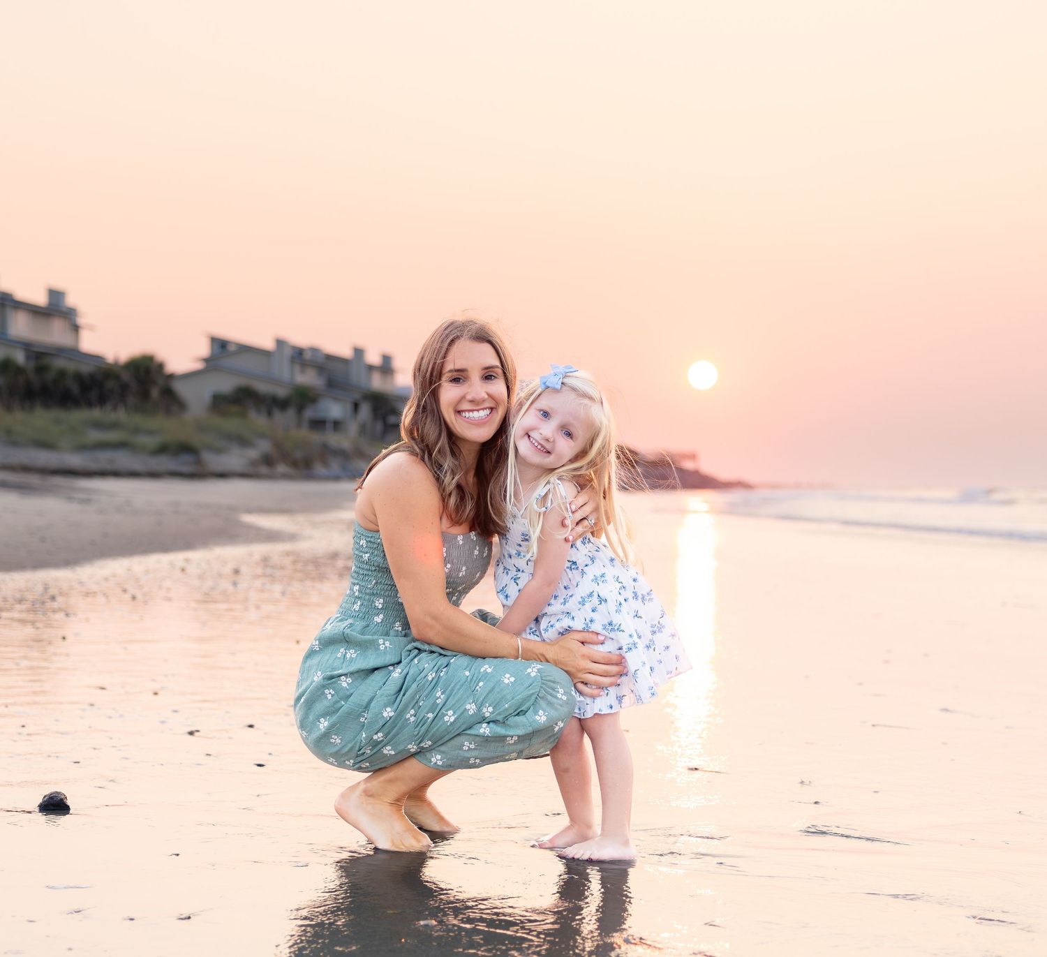 Woman and young girl in matching dresses smiling on a beach at sunset with ocean waves behind them.