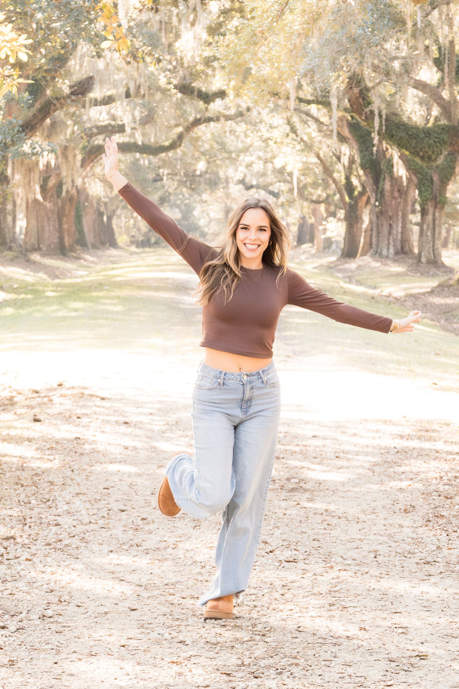 Woman in brown crop top and jeans balancing on one leg with arms outstretched on a sunlit tree-lined path.