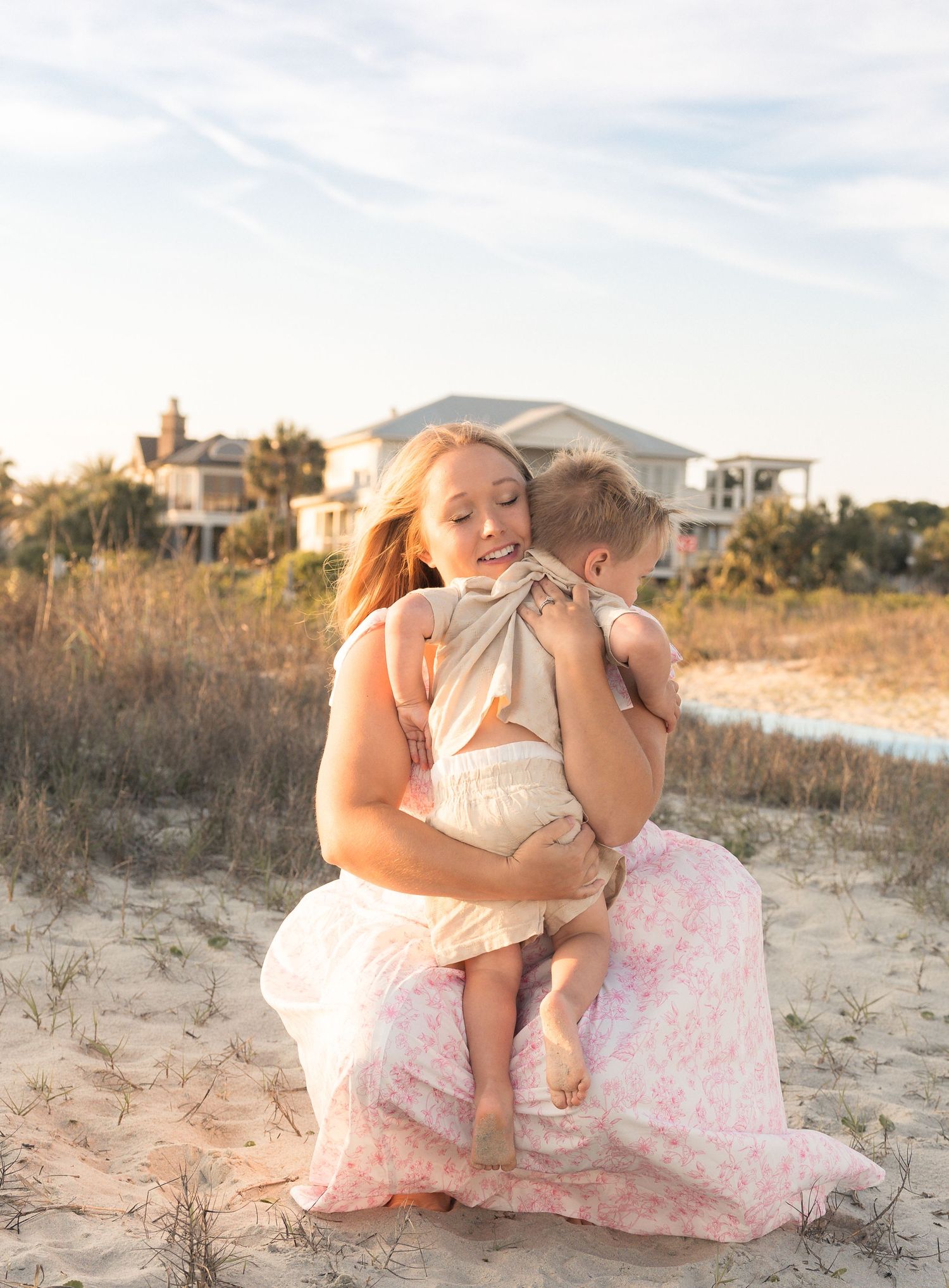 A mother kneels on sandy beach holding her toddler close, wearing a pink floral maxi dress at golden hour.