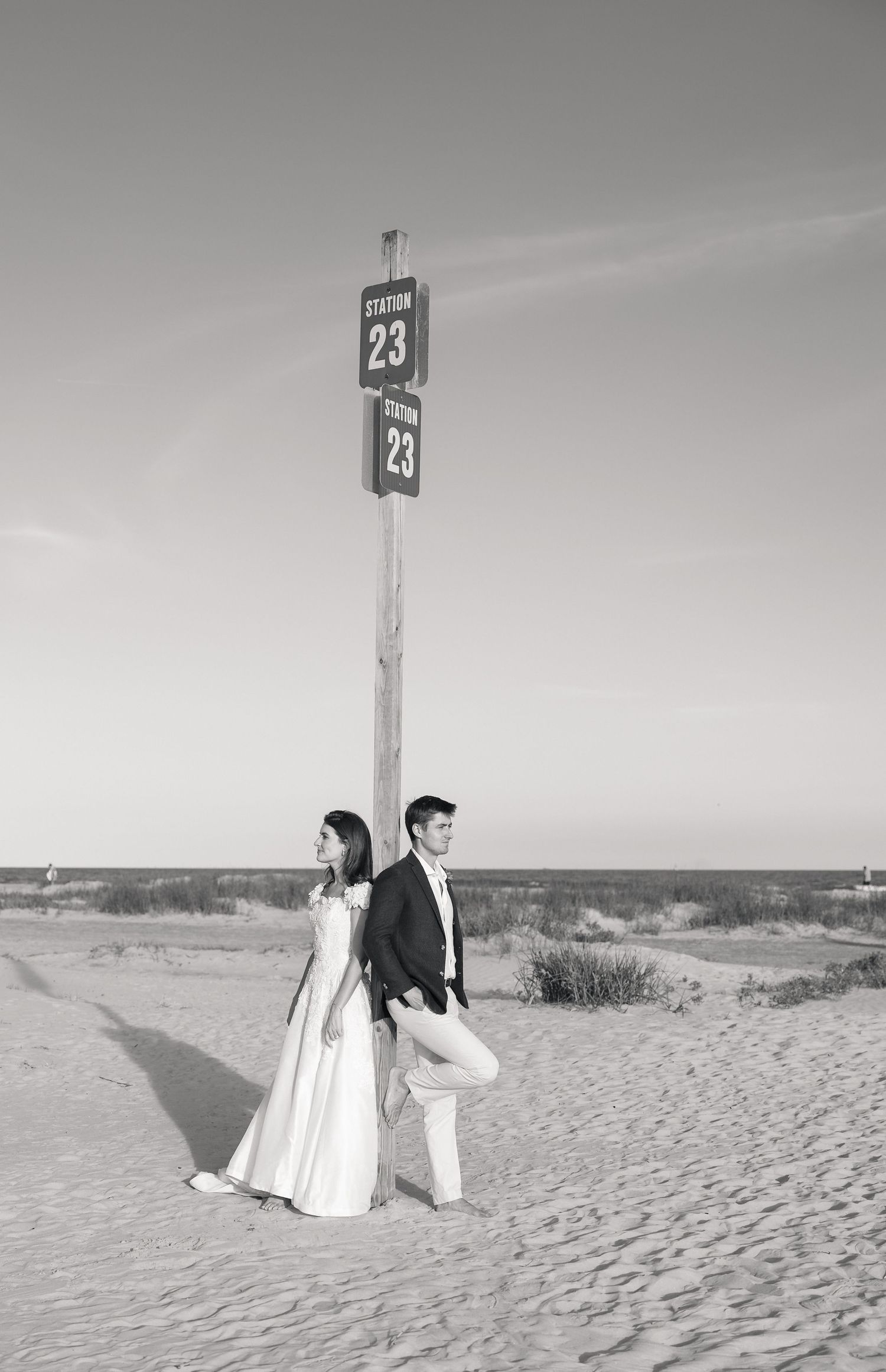 Bride and groom stand back-to-back at beach Station 23 sign on sandy shore in black and white wedding photo.
