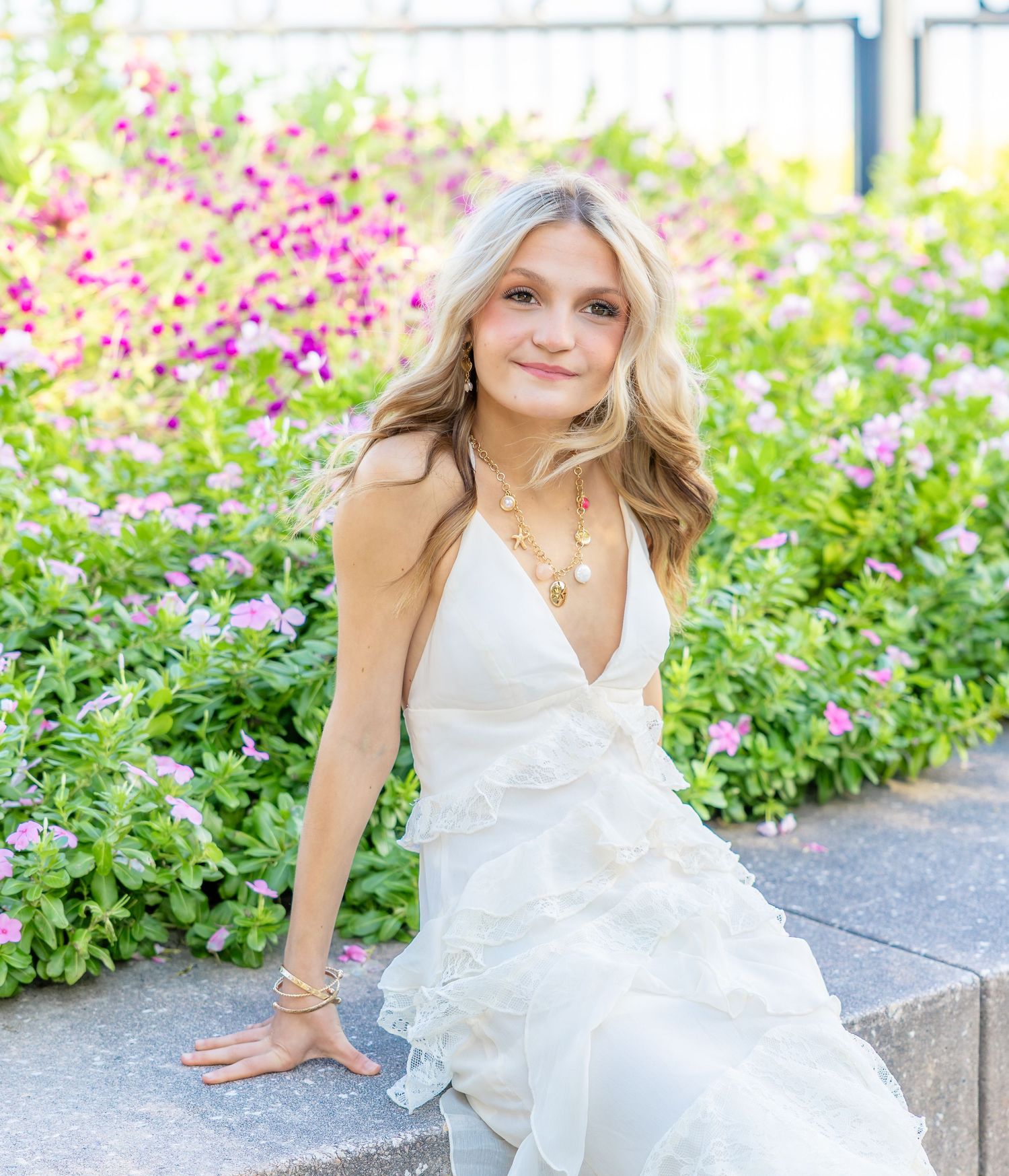Young woman with long wavy blonde hair in a white ruffled dress sits on a stone ledge beside blooming pink and purple flowers.