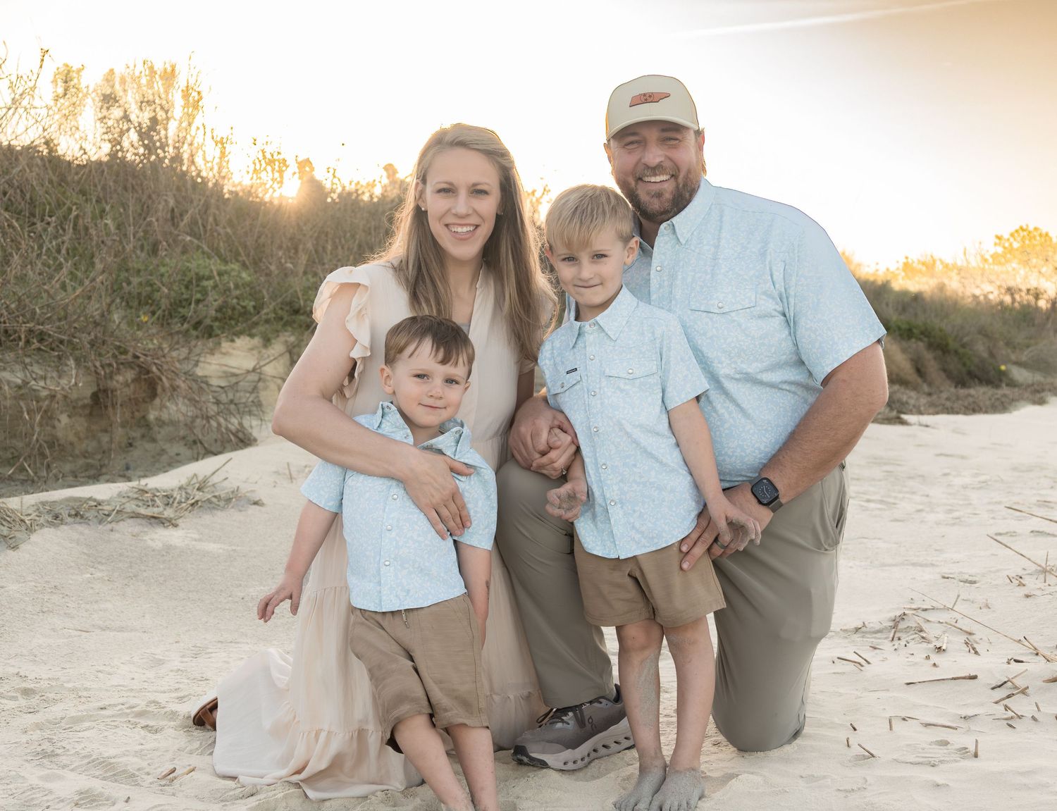 Family of four smiling together on a sandy beach at golden hour, with dunes and trees in the background.