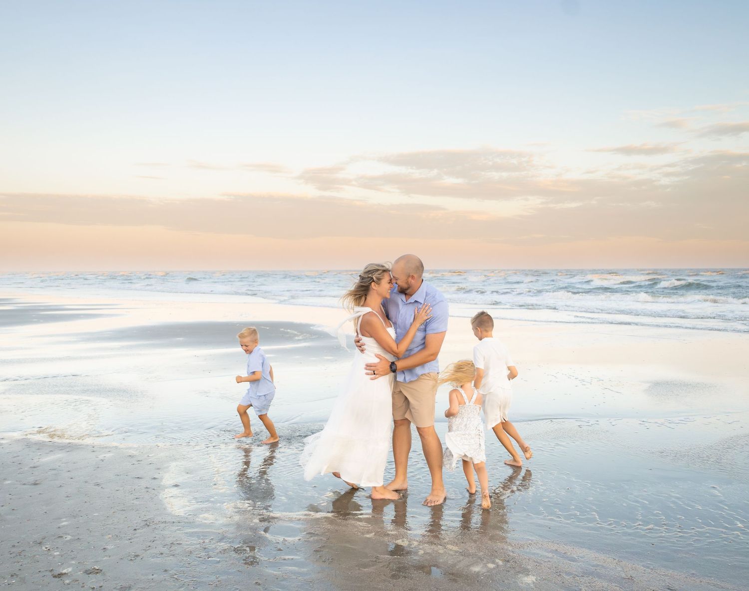Family in white outfits embracing on a beach at sunset with children playing in the waves around them.