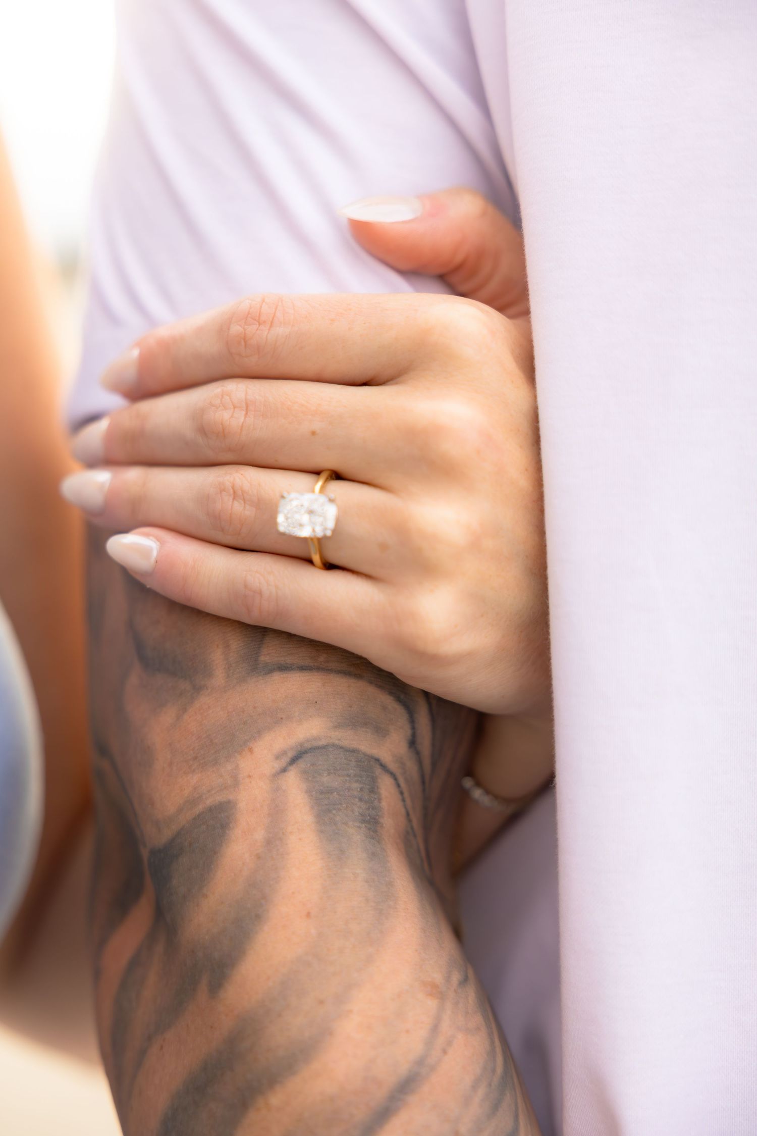 Close-up of a woman's hand wearing a gold solitaire diamond engagement ring resting on a tattooed arm.