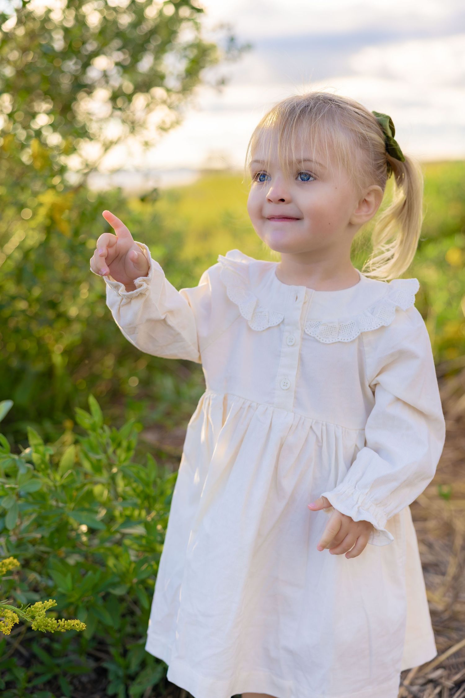 Toddler girl in a cream ruffle-collar dress pointing outdoors in a sunlit green field with her blonde hair in a ponytail.